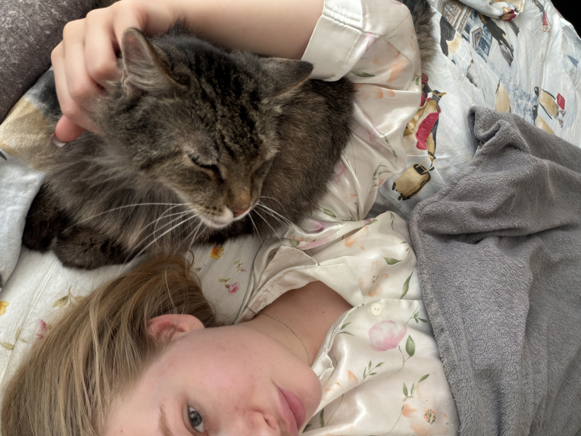 A woman is lying down, petting a brown and black tabby cat. The woman has light skin and blonde hair. She is wearing a light-colored floral pajama top. The cat is mostly brown and black with white whiskers. They are lying on a bed with floral-patterned bedding and a gray blanket. The scene is indoors. The location is Засечное, Russia.