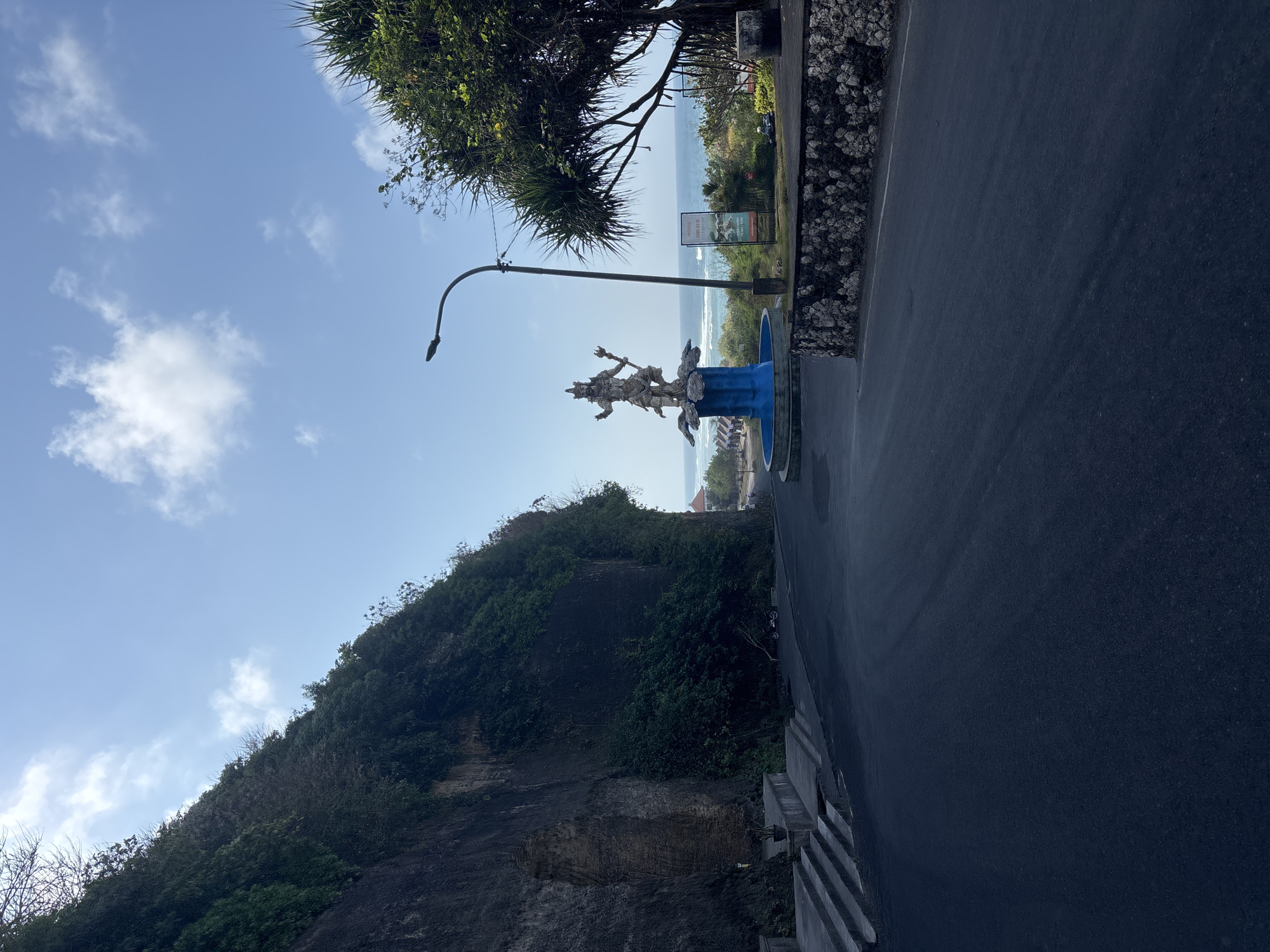 The image shows a statue of a deity standing on a blue fountain. The statue is located in Kuta Selatan, Indonesia, with a road in the foreground and a cliff face on the left. The ocean is visible in the background, with waves crashing on the shore. The sky is blue with some clouds. A lamppost stands near the statue. The scene appears to be a tourist attraction.