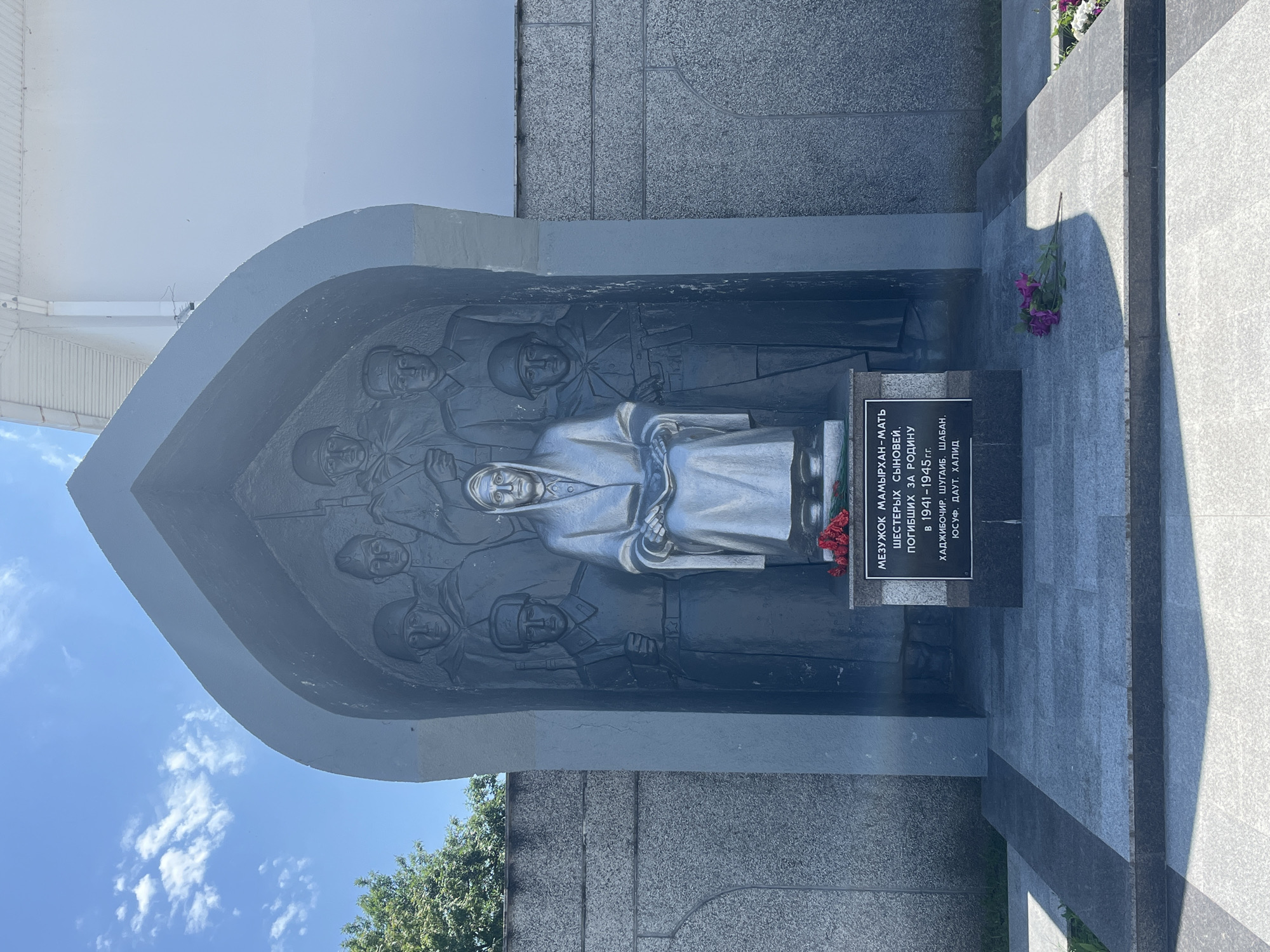 The image depicts a memorial in the city of Takhtamukai, Russia. The central subject is a sculpture group within a recessed, arched structure. The sculpture features a central seated female figure draped in fabric, holding what appears to be a book or scroll. Surrounding her are five standing figures of soldiers in military attire, some holding rifles. The background of the recessed area is textured, resembling bas-relief. Below the sculpture, a dark plaque is affixed to a stone base, bearing inscriptions in an unidentifiable script, and is adorned with red flowers. In front of the base, on a stone-paved area, are more flowers, some purple and some white. The memorial is set against a stone wall on the left, and a white building with visible architectural details on the right. The sky above is blue with scattered white clouds. The overall scene is solemn and commemorative.