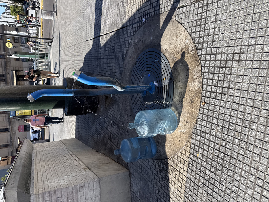 A public drinking fountain with two blue spouts dispenses water, filling two large plastic bottles. The fountain sits on a circular, tiled base with a drain. People stand in the background. The scene is cast in harsh sunlight, creating strong shadows. The surrounding area is paved with small square tiles.