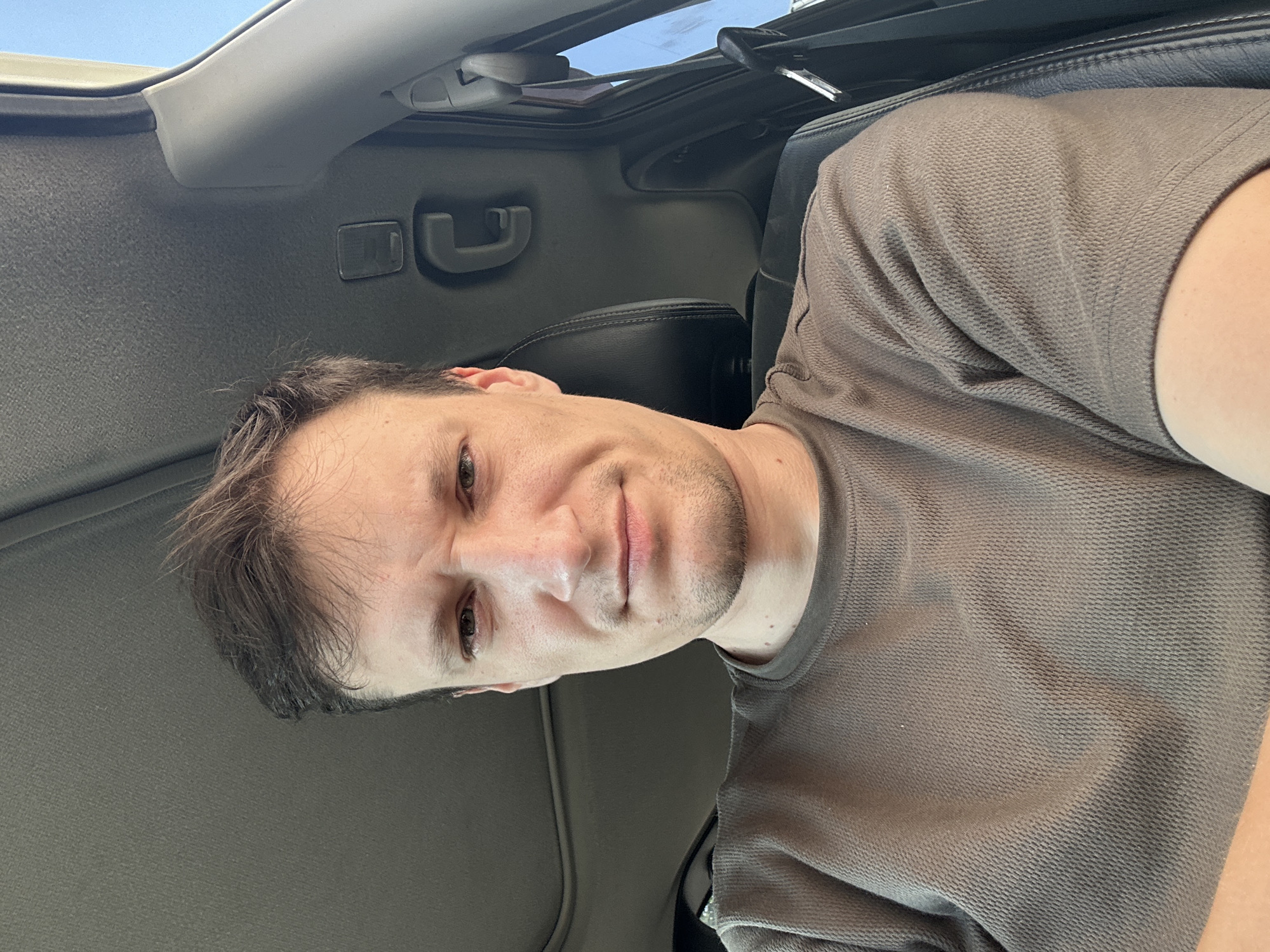 A selfie of a man in a vehicle, with his face positioned centrally in the frame. He is wearing a brown, long-sleeved shirt with a textured fabric. The man has short, dark brown hair and light-colored eyes. His expression is neutral, with slightly furrowed brows. The interior of the vehicle is visible, including a dark headliner, a sunroof, a black leather seat, and a portion of the car door with a black seatbelt buckle. The lighting suggests it is daytime, with light entering from the side window. The image does not contain any identifiable landmarks or visual cues that would indicate the specific location within Shatava, Ukraine.