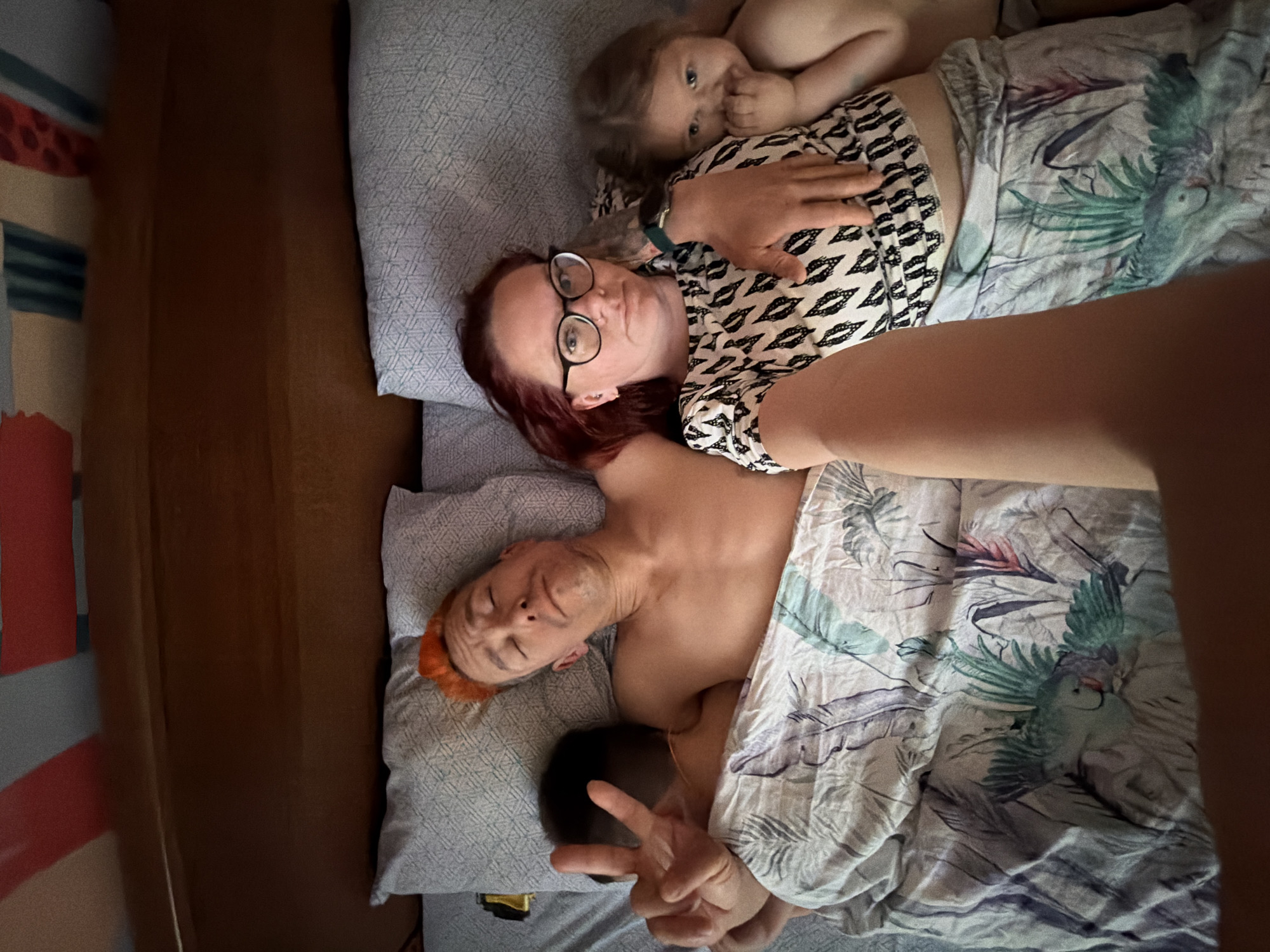 A selfie photo taken in a bedroom. A woman with red hair and glasses is lying on a bed, facing the camera. A small child is lying on her chest. Another person with orange hair is lying partially under the covers, facing away from the camera, giving the peace sign. The bedding has a tropical bird pattern. The location is inferred to be in the city of Chaikovsky, Russia.