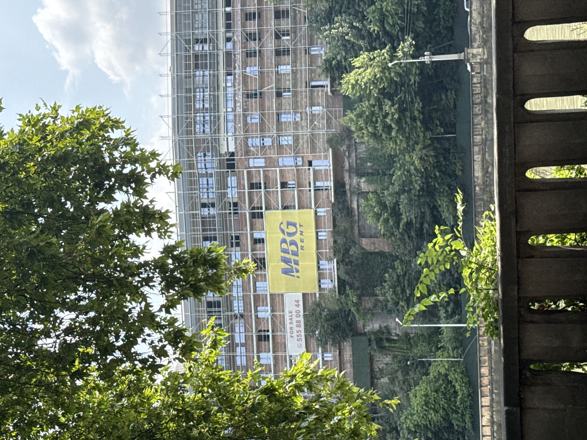 This is a medium-shot, eye-level photograph of a multi-story brick building under construction, viewed from across a body of water and behind a concrete barrier. The building is partially obscured by the leaves of a large tree in the foreground. Scaffolding covers much of the building's facade, and windows are visible in rows. Two large banners are hung on the building: one yellow banner with the text "MBG RENT" and a smaller white banner that reads "FOR SALE" and displays a phone number "555 88 00 44". The building is situated on a steep, tree-covered embankment. In the middle ground, beyond the water, two streetlights are visible, and behind them, a dense forest covers the hills. The sky is partly cloudy. The location is the Left Bank of the Mtkvari River in Tbilisi, Georgia, specifically near the Bridge of Peace.