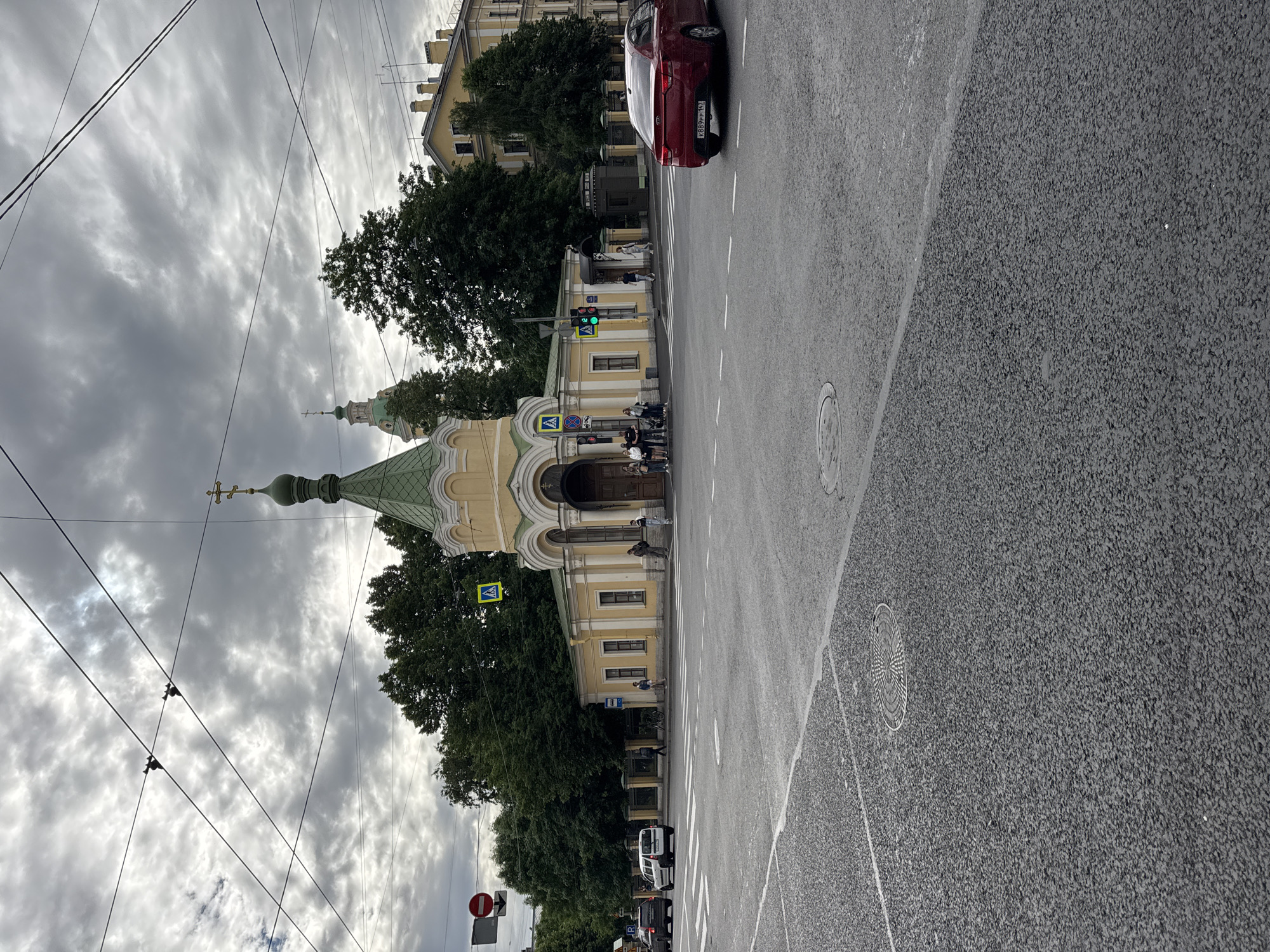 The image depicts a street scene in Saint Petersburg, Russia, on an overcast day. The focal point is a light yellow chapel with a green, conical roof topped with a cross. The chapel is situated on a corner, bordered by trees and a yellow building with a similar architectural style. Several people are visible standing near the chapel entrance and on the sidewalk.
In the foreground, a wide asphalt road with white pedestrian crossings dominates the lower half of the image. A red sedan is driving away from the viewer on the right side of the road. Traffic lights are visible, showing a green light. Overhead, a complex network of electrical wires crisscrosses the sky, suggesting the presence of trams or trolleybuses. The sky is heavily clouded, with grey tones dominating.
Other elements in the scene include road signs, a bus stop shelter, and a bicycle parked near the chapel. The overall impression is of a typical urban environment with both historical architecture and modern infrastructure.