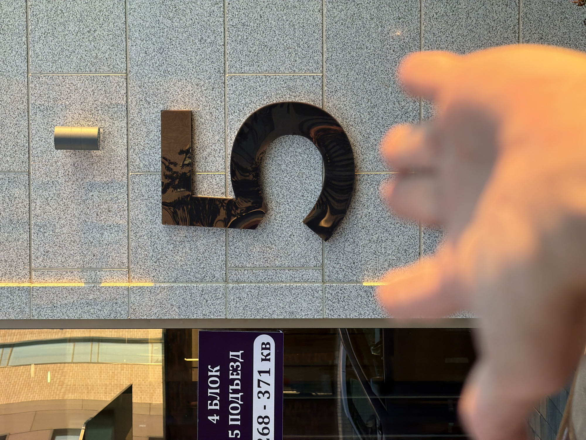 The image depicts a close-up view of a building facade with a prominent number "5" mounted on it. The number is large, glossy black, and appears to be made of metal or plastic, casting a reflection. Above the number "5" is a cylindrical gray light fixture. To the left of the number, a purple sign with white text is visible, reading "4 БЛОК 5 ПОДЪЕЗД 268 - 371 КВ". This text is in Russian and likely indicates building information such as block number, entrance, and possibly apartment range. The facade is constructed of light-colored granite tiles. In the foreground, a blurry human hand, illuminated by warm, golden sunlight, is extended towards the camera, obscuring the bottom portion of the number "5". The background includes reflections on a glass-fronted building, showing architectural elements and what appears to be brickwork. The scene suggests an outdoor urban environment, likely on a street in Astana, Kazakhstan, given the context provided. The lighting indicates it is either sunrise or sunset.