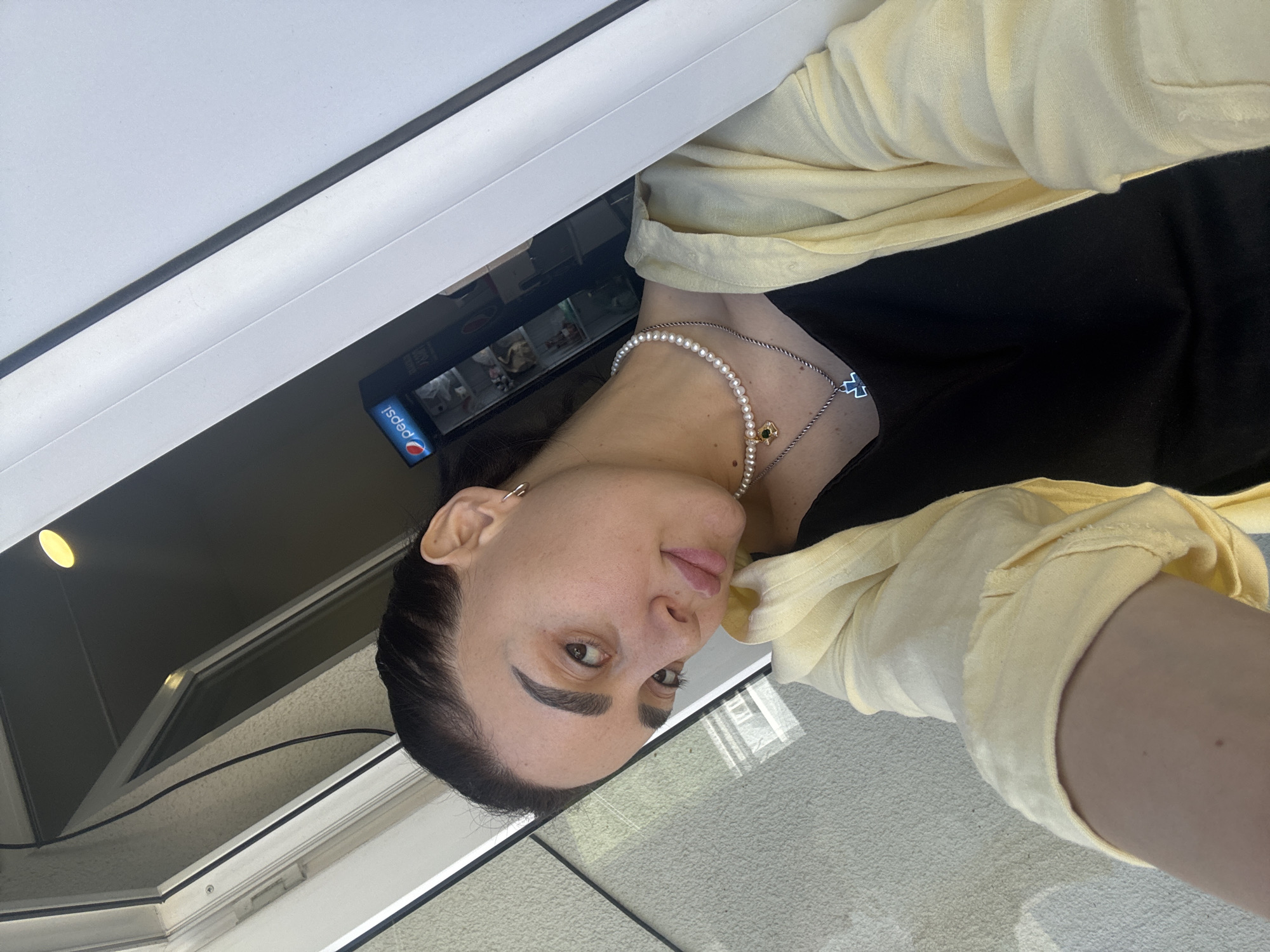 A woman is positioned inside a building, likely in Tbilisi, Georgia. She is wearing a black top and a loosely-buttoned, light yellow shirt. A pearl necklace with a small pendant is visible around her neck. She is standing near a window, with a vending machine containing Pepsi products visible behind her. The surroundings appear to be modern and indoors.