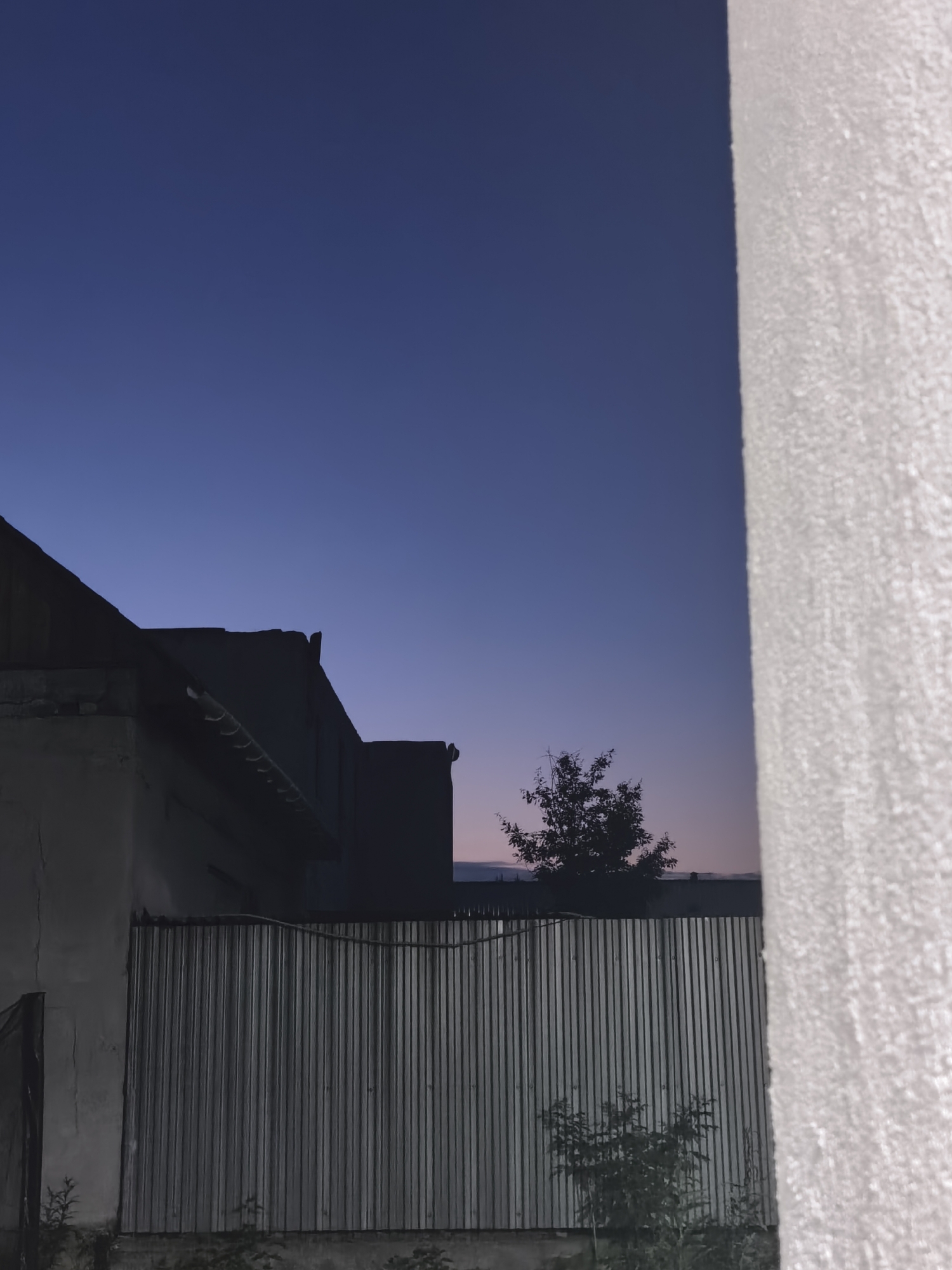 The image depicts a twilight scene in what appears to be a residential or semi-rural area. A large, rough-textured white wall dominates the right side of the frame. To the left, silhouetted against a deep blue sky transitioning to pale pink at the horizon, are the rooftops of buildings and a corrugated metal fence. A lone tree is visible in the middle ground, its branches stark against the fading light. The overall impression is one of quiet dusk, with a focus on architectural and natural elements. The location is identified as Белоярская Застава, Russia.