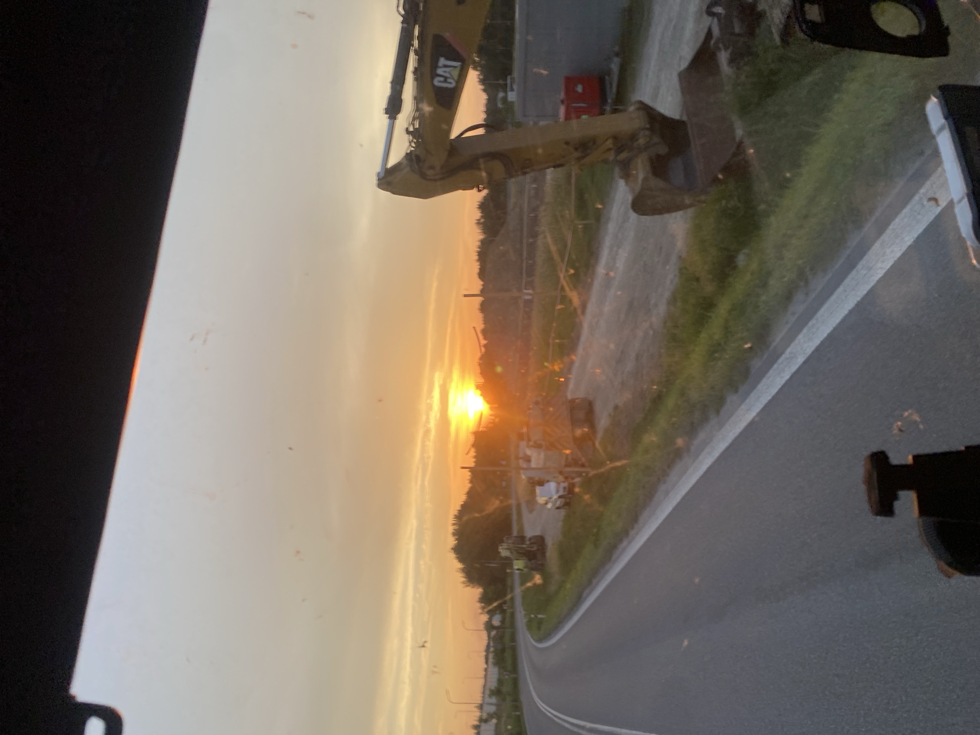 The image shows a construction site at sunset in Podłęże, Poland. A yellow CAT excavator is the prominent object in the foreground, with its arm extended. Beyond the excavator, a road curves to the right, bordered by a grassy verge and trees. The sky is filled with the warm hues of sunset, casting a golden light over the scene. A few small vehicles are visible in the distance. The overall impression is one of industrial activity at the end of the day. The time of day is clearly sunset, indicated by the low angle of the sun and the warm colors in the sky. The weather appears to be clear. The location is specified as Podłęże, Poland. No people are visible in the image. The text "CAT" is visible on the excavator.