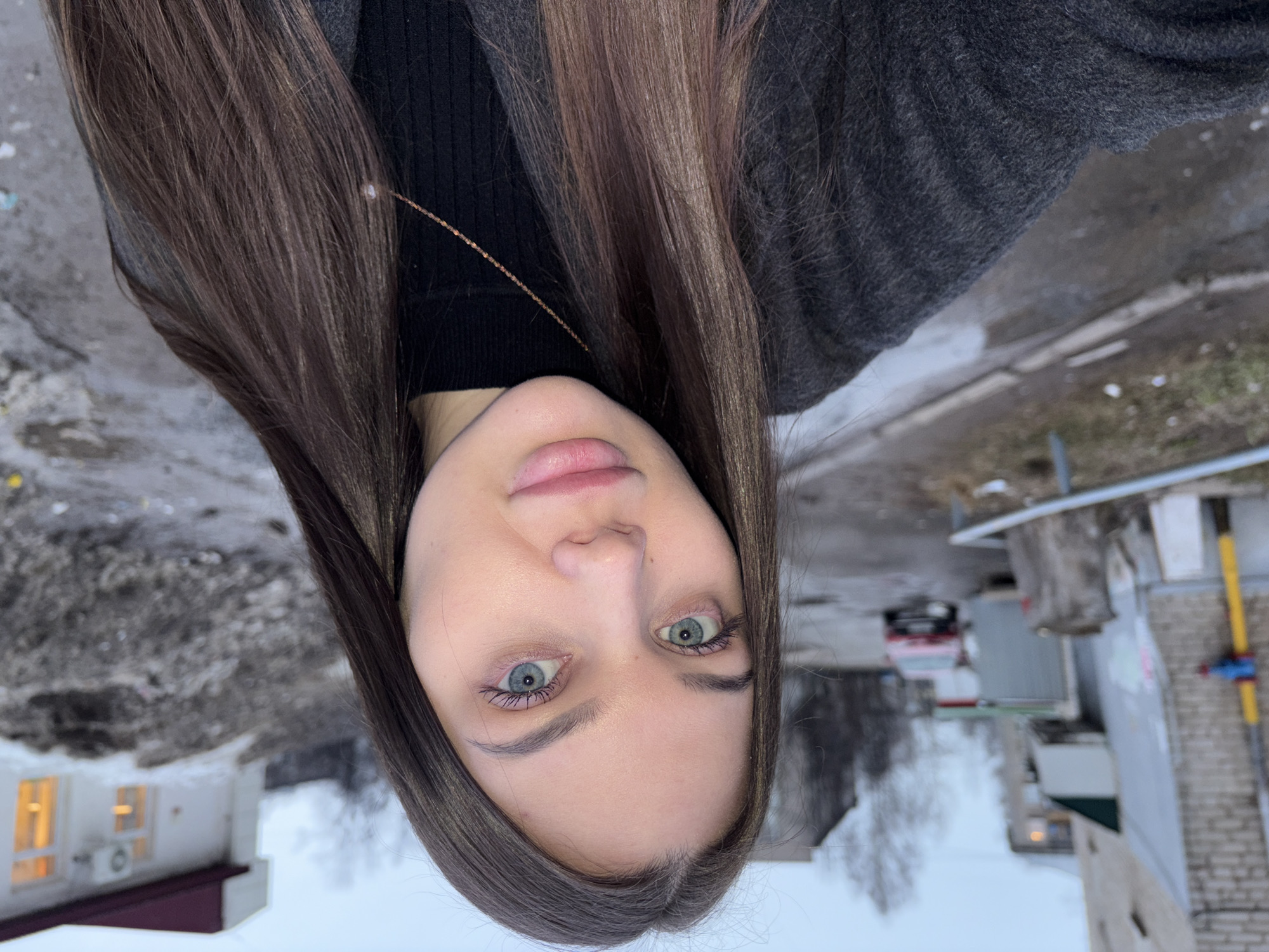A selfie image shows a light-skinned woman with long brown hair, light blue eyes, and wearing a dark sweater and gray jacket. The background appears to be a street scene in Chaikovsky, Russia, with buildings, snow, and leafless trees visible. The image is rotated.