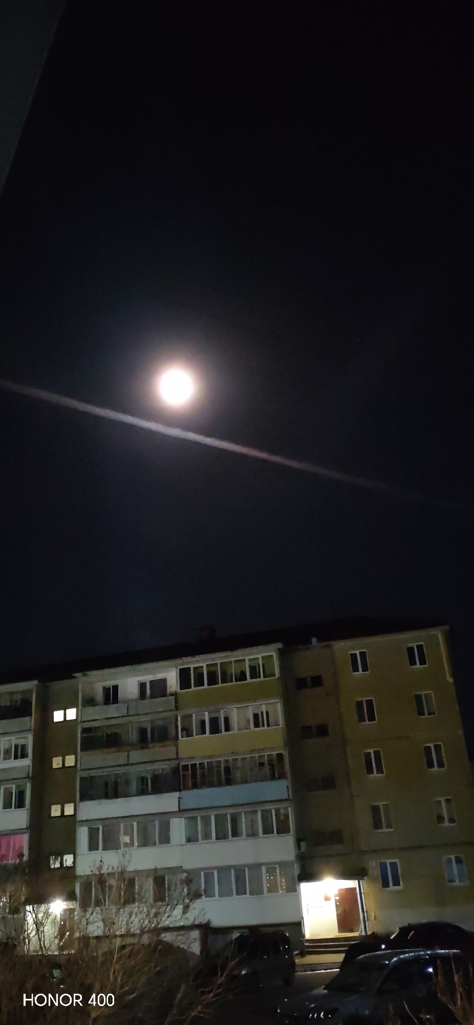 The image shows a multi-story residential building in Noviy Urengoy, Russia, at night. The sky is dark with a bright moon and a contrail across it. The building has multiple floors with illuminated windows. There are trees in the foreground and parked cars. The image was taken with an HONOR 400 mobile phone.