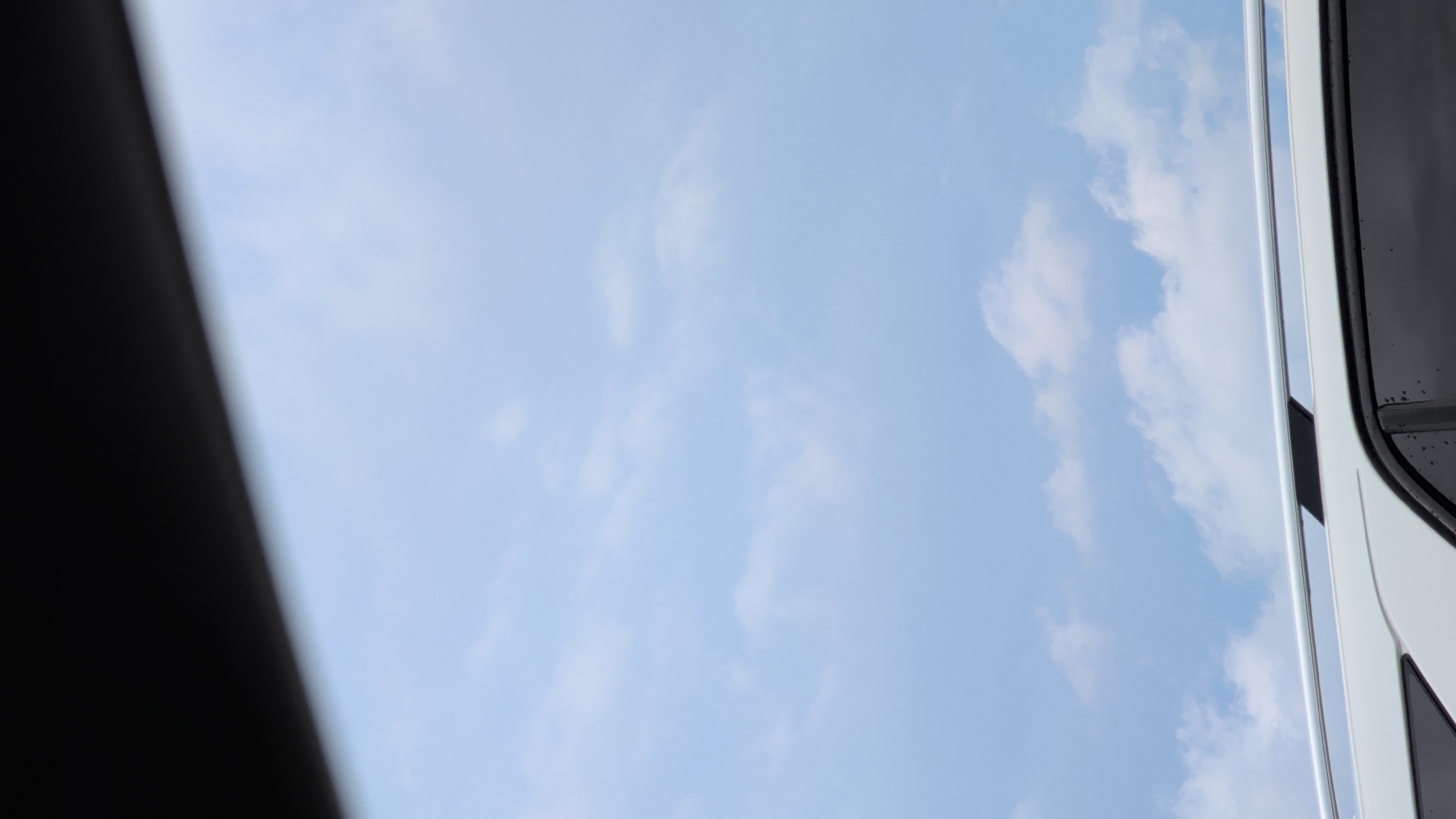 The image shows a sky with scattered clouds, seen from the perspective of inside a vehicle. The top of a white vehicle, including its roof rack and a portion of its window, is visible in the foreground. The scene appears to be set outdoors, possibly in the city of Dorchester, Canada, given the information provided. There are no people or specific actions depicted.
