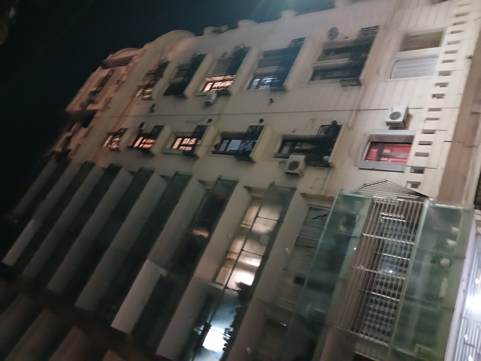 The image shows a multi-story building facade at night. The building has a light-colored exterior with rows of windows, some with awnings or balconies. Many windows are illuminated from within. Air conditioning units are mounted on the exterior walls below some windows. A glass-enclosed structure is visible at the lower right of the image. The angle of the shot is looking upwards at the building, implying the picture was taken from street level. The location is likely in Buenos Aires, Argentina, based on the architectural style and urban setting.