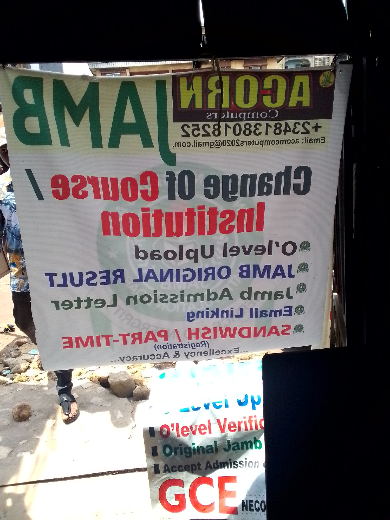 In Nkpor, Nigeria, a vibrant banner, likely advertising educational services, hangs from a dark, overhead structure. The banner, predominantly white with bold green and red lettering, is oriented to be read upside down. Dominating the top of the banner is the word "ACORN" in large, yellow and green lettering, with the word "COMPUTERS" in smaller white letters below it.
The banner lists various services and requirements. The phrase "CHANGE OF COURSE" is prominently displayed in red, followed by "INSTITUTIONAL" in red. Further down, "O' LEVEL" and "O'LEVEL VERIFICATION" are visible in green and red respectively. There's also mention of "ORIGINAL JAMB" and "ACCEPT ADMISSION". Other services include "JAMB ORIGINAL RESULT", "JAMB LETTER", "SANDWISH", and "PART-TIME". The text "registration" and "Accuracy" are also present. A phone number, "+2348038184838", and an email address, "acorncomputers@slim.com", are also listed.
A person, partially visible on the left side of the image, is wearing a blue and white patterned shirt and dark pants with sandals. Their left leg is bent, and their foot is resting on the ground. The environment appears to be an outdoor setting with some loose soil and stones on the ground. In the lower right portion of the image, a black rectangle obscures a part of the scene.