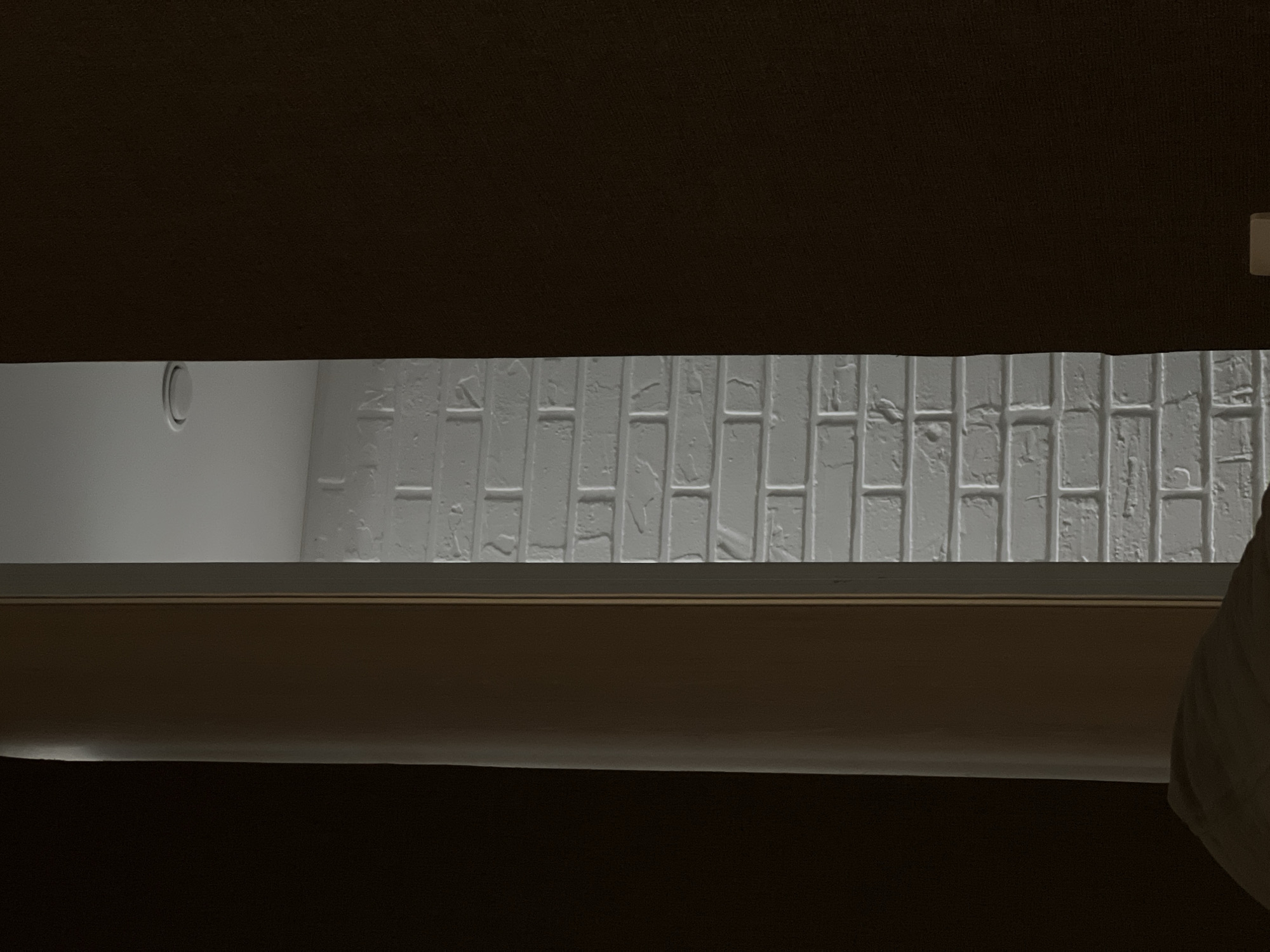 The image appears to be a view through a partially opened window or a similar opening. The foreground and background are dominated by dark brown surfaces, likely part of a wall or furniture. Through the opening, a white brick wall is visible, along with a section of a white wall with a circular vent. The location and specific city are unknown.
