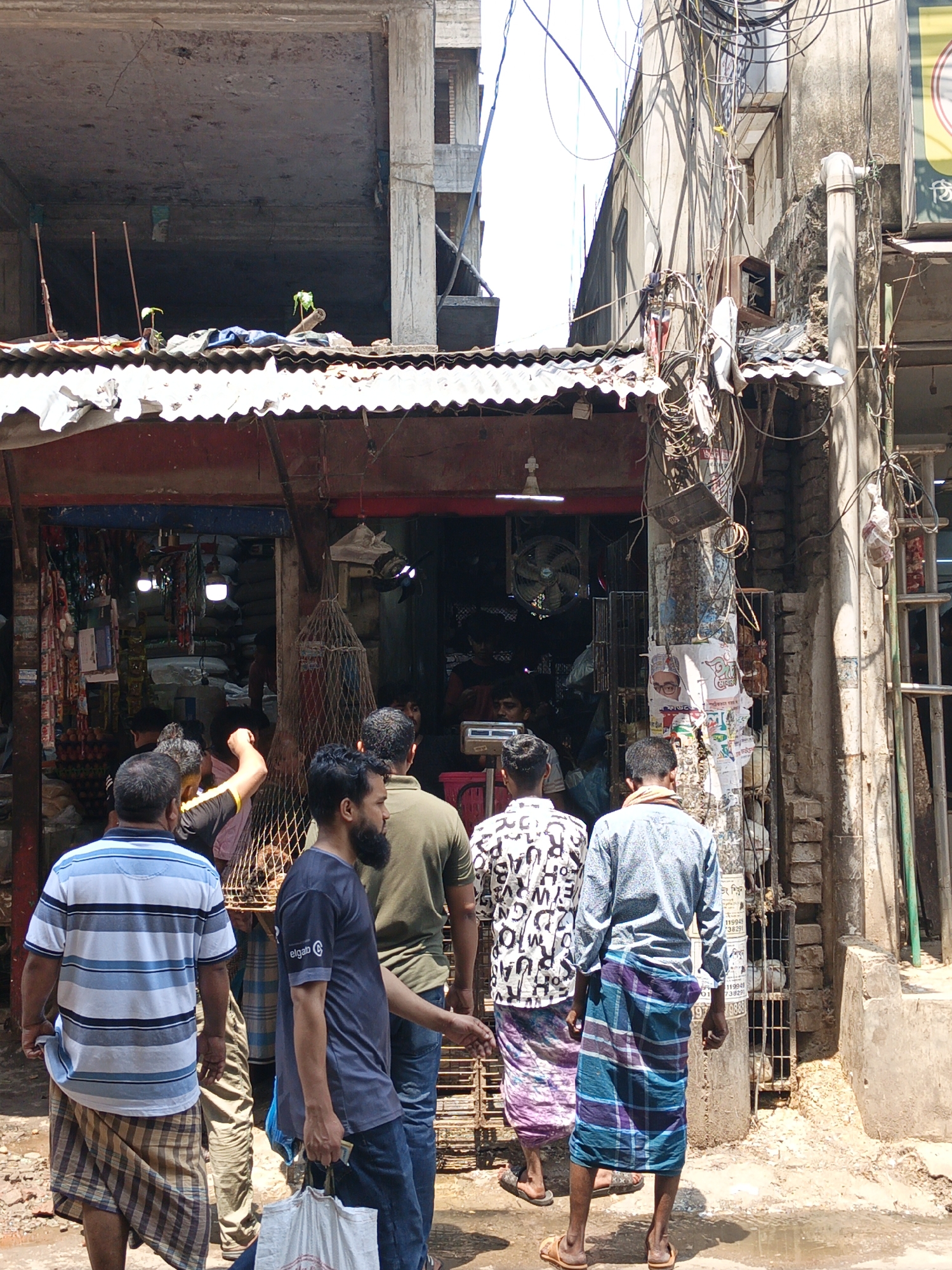 The image depicts a street scene in Dhaka, Bangladesh. In the foreground, several men are standing and walking on a street toward a small shop under a dilapidated awning. Most of the men are wearing lungis and shirts. An unfinished concrete building is overhead. The shop is dimly lit, displaying merchandise and attended by figures inside. Utility wires are densely tangled above the shop and along an adjacent building. The environment is urban, with signs of age, wear, and activity. The overall impression is of a busy, everyday scene in a densely populated area.