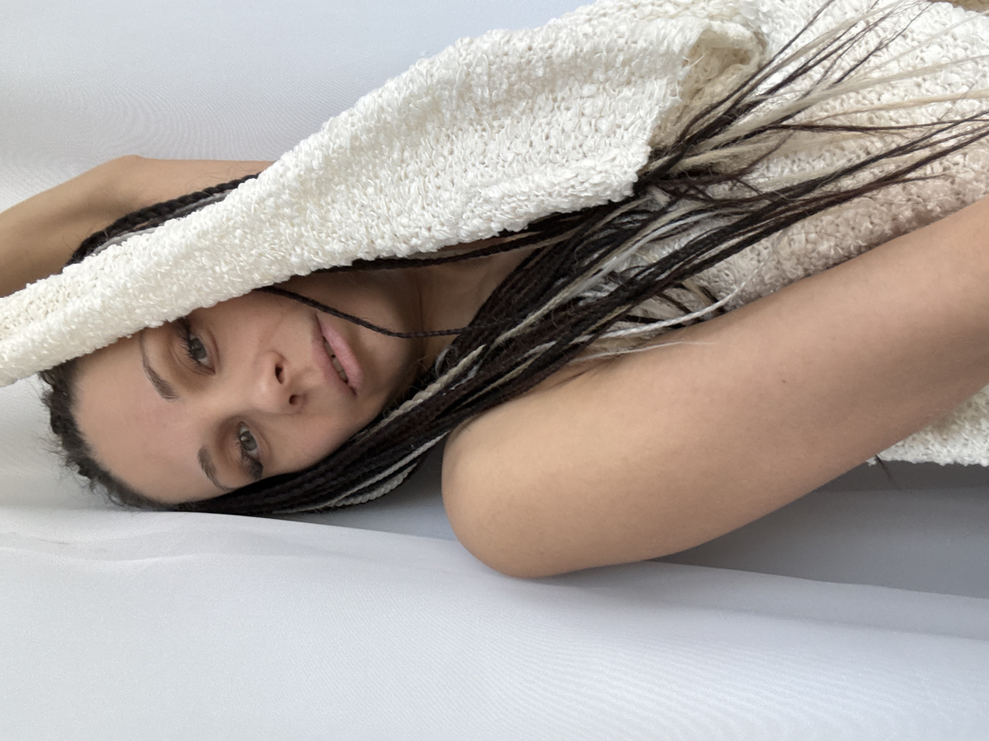 A woman lies on a white surface, possibly fabric, partially covered by a cream-colored, loosely woven garment. Her head rests on the surface, and her gaze is directed towards the viewer. She has light skin, light-colored eyes, and braided hair with a mix of dark and light strands. One strand of hair is visible across her face near her mouth. Part of her shoulder and arm are visible in the foreground. The background appears to be a continuation of the white surface. There are no indications of the image being taken in Rusanovo, Russia, or any other specific location.