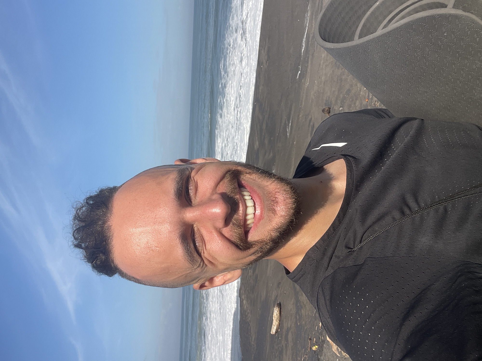 A man is smiling into the camera with a beach and ocean behind him. He is wearing a black shirt and standing on black sand. A yoga mat is rolled up in the background. The sky is blue. The location is Kediri, Indonesia.