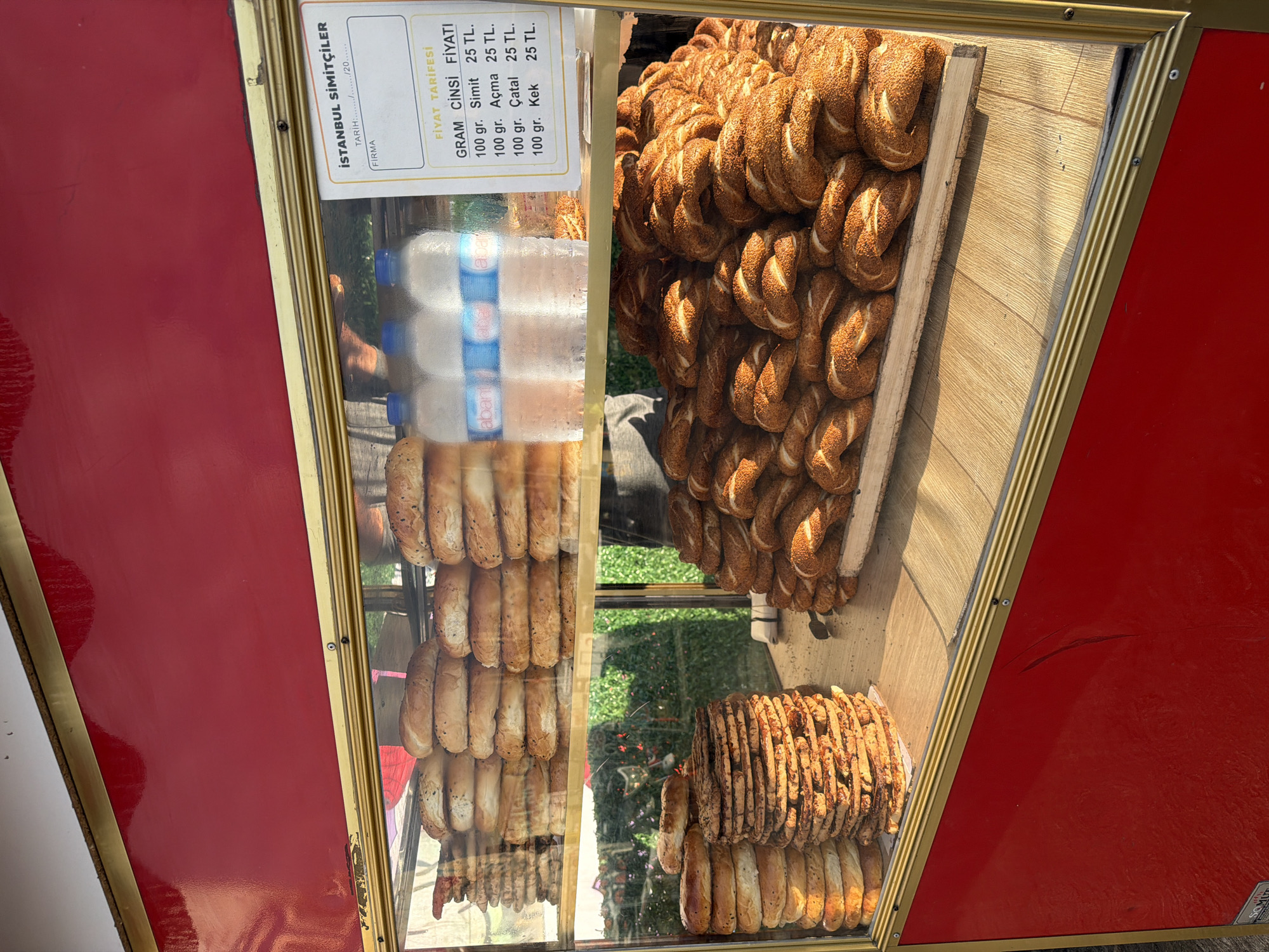 The image shows a display of baked goods, likely from a street vendor in Beyoğlu, Turkey. The items are arranged in a glass-fronted case with a red exterior. Piles of simit, a circular bread with sesame seeds, are prominently featured. There are also stacks of other types of bread and flatbreads. Bottles of water are visible behind the bread. A price list is displayed on the right side of the case, listing prices for simit, açma (a type of Turkish pastry), and kek (cake) by weight. The environment appears to be outdoors, with greenery visible through the glass in the background. The lighting suggests it is daytime.
