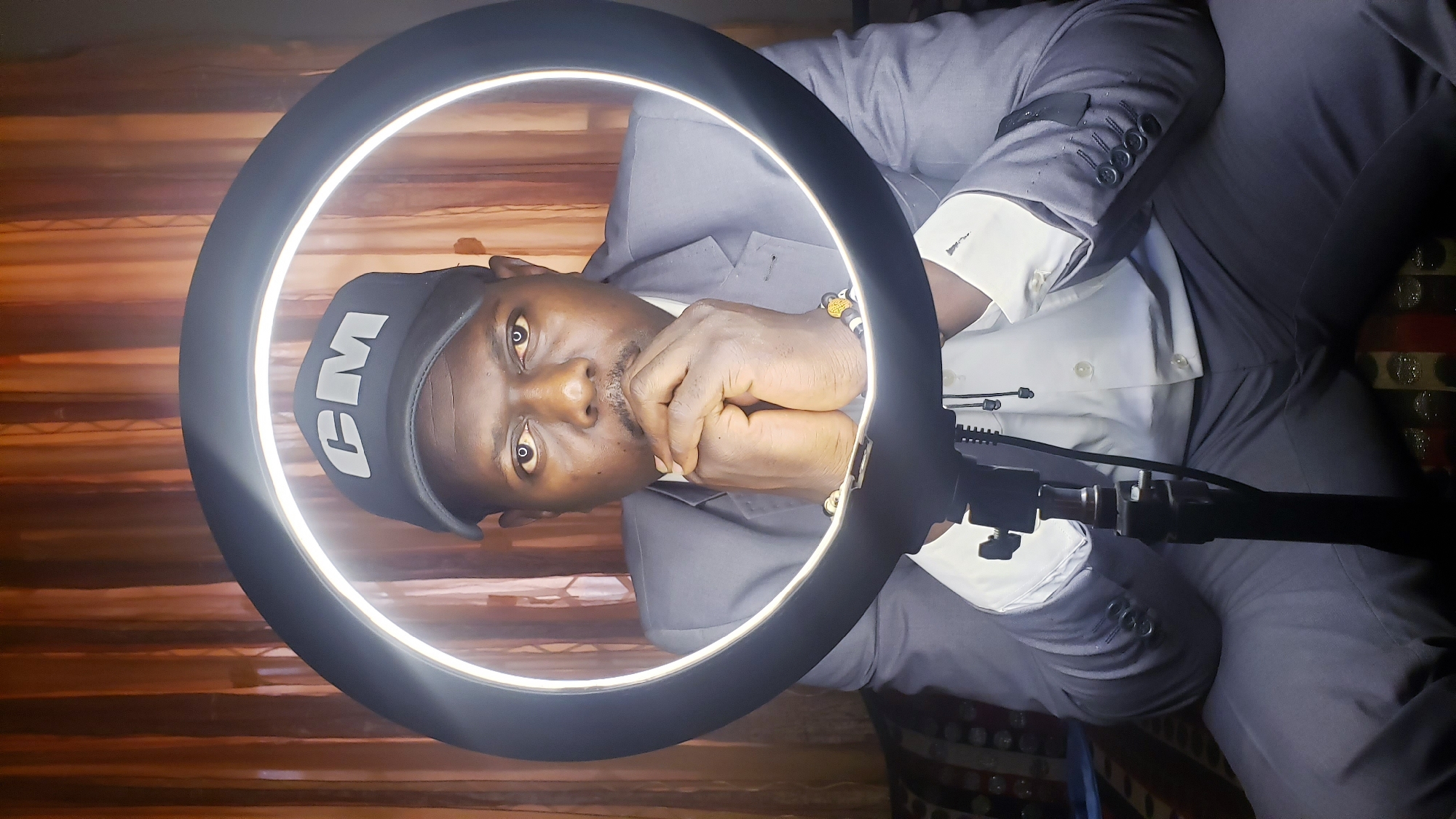 The image features a dark-skinned man, seemingly indoors in Sabon Tasha, Nigeria, with a bright ring light illuminating his face. He is looking directly into the camera with a neutral, thoughtful expression, his hands clasped near his chin.
The man is dressed in a grey suit jacket over a white collared shirt. He wears a black cap with the white letters "CM" clearly visible on the front. A multi-colored beaded bracelet is on his right wrist. The ring light frames his head and shoulders, creating a bright, even illumination and distinctive circular reflections in his eyes.
The background is dimly lit compared to the subject. It features textures that suggest either wooden wall paneling or a patterned curtain. To the right side, a section of a patterned cushion or seat is visible. The setup suggests a deliberate pose for photography or video, utilizing the ring light for professional lighting.