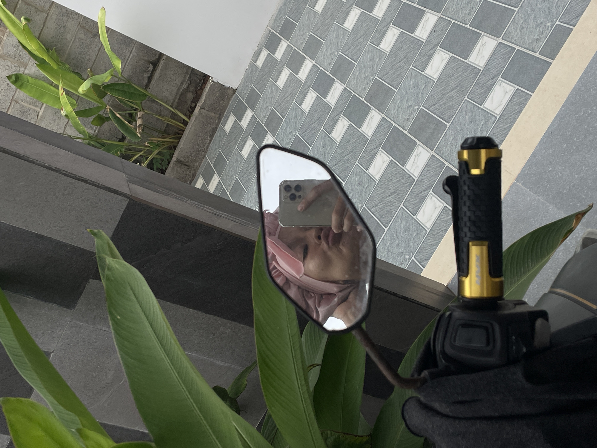 The scene appears to be an outdoor area in Phu Thuy Ward, Vietnam, based on the tiled flooring and architectural style. A motorcycle handlebar with a black grip and gold accents is visible on the right. To the left of the handlebar, vibrant green plants with long, broad leaves are present. The motorcycle's side mirror reflects a person holding a smartphone, capturing a selfie. The flooring consists of grey tiles with white geometric patterns. In the upper-left, there are additional plants growing near a wall.