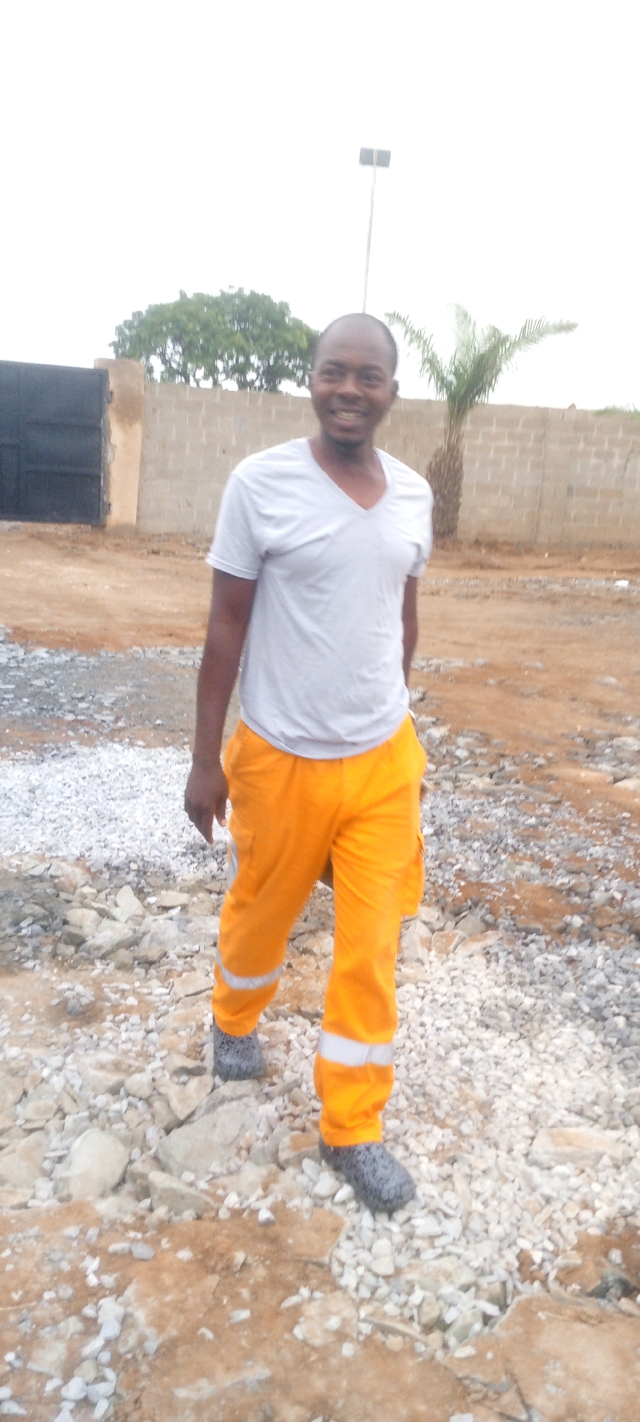 A Black man is standing on a construction site. He is wearing a light gray V-neck t-shirt and bright orange work pants with reflective white stripes around the calves. He is also wearing dark gray, possibly knitted, foot coverings that extend up to his ankles. His left hand is visible at his side, and he is looking towards the camera with a slight smile. Behind him, a beige brick wall runs horizontally, with a dark metal gate on the left. A tall pole with a light fixture is visible in the background, as is a palm tree. The ground is covered with gravel and dirt, indicative of an outdoor construction or undeveloped area. The sky is overcast.