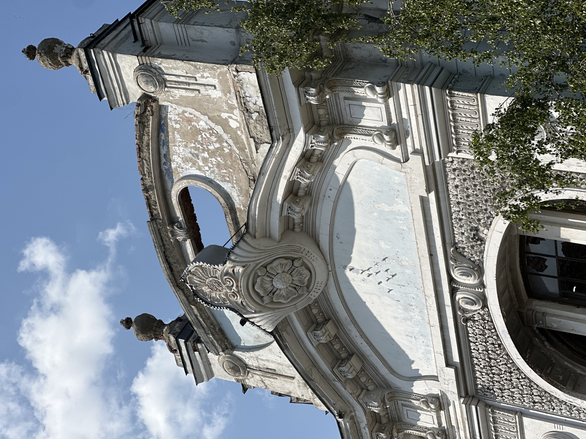 This is a low-angle, medium shot of the upper facade of a decaying ornate building, likely part of the former Biysk City People's House (now the Altai State Institute of Culture and Arts). The sky is clear blue with scattered white clouds. The building's architecture features classical elements, including decorative stucco work.
The facade itself shows significant disrepair. Large sections of white and pale blue paint are peeling off, revealing older layers and crumbling plaster underneath. The roofline is damaged, with exposed brickwork visible in places. A prominent circular opening is present in the upper part of the facade.
Elaborate decorative elements are visible, including a large floral motif with radiating petals and a detailed frieze of smaller floral designs below it. The architectural ornamentation includes scrolls, cornices, pilasters, and finials.
To the right of the building, the branches of a green, leafy tree are partially obscuring the view. No people are present in the image, and there are no obvious signs of ongoing activity. The overall impression is one of faded grandeur and neglect.