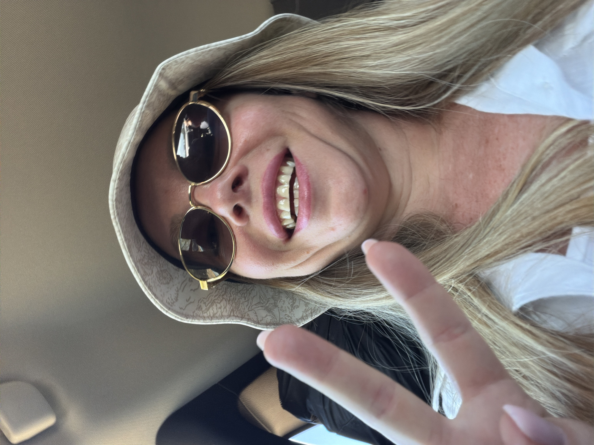 In a selfie taken from a low angle, a blonde woman wearing a white bucket hat and sunglasses smiles broadly. Her mouth is open, revealing her teeth, and she is holding up two fingers in a peace sign. She appears to be in a car, with the ceiling and a black seat visible in the background. The woman is looking directly at the camera, and her blonde hair falls around her shoulders. The lighting suggests it is daytime. There is no specific information in the image to indicate the location as Άγιοι Απόστολοι, Greece.