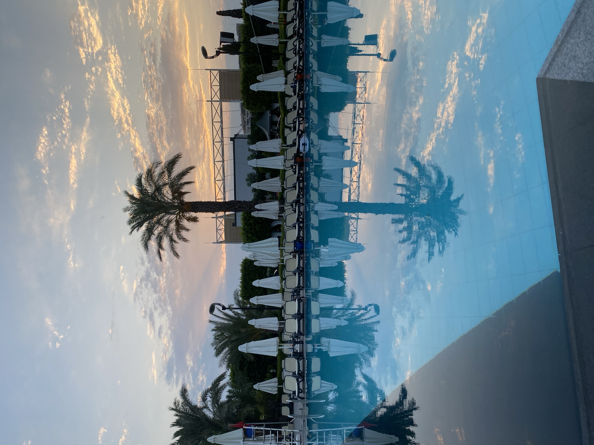 A vertical, eye-level shot captures a swimming pool and its surroundings during what appears to be sunrise or sunset in Aksu, Turkey. The sky is filled with soft, wispy clouds tinged with orange and yellow hues, with a pale blue gradient above. The water in the foreground is calm and reflects the sky and surrounding elements.
Lined up neatly along the edge of the pool are numerous white, closed beach umbrellas and lounge chairs with beige cushions. A lifeguard chair with a red flag is visible on the left. Palm trees and other greenery form a backdrop behind the chairs and umbrellas. In the distance, structures with white horizontal beams and some buildings are discernible, along with a tall light fixture. The reflection in the pool mirrors the sky, palm trees, umbrellas, and chairs with striking clarity, divided by a diagonal line where the dark pool edge meets the lighter water.