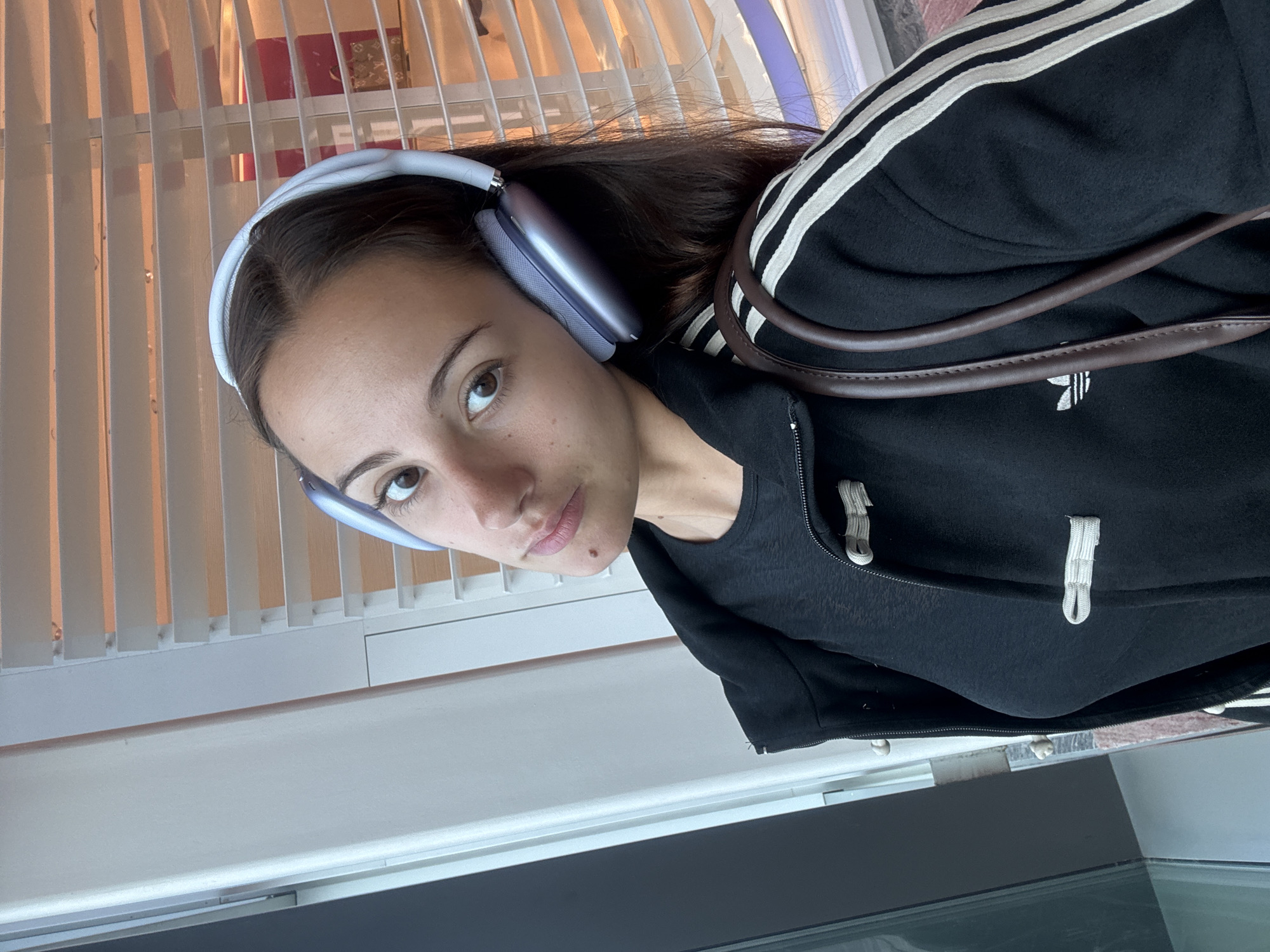 A young woman with brown hair and light brown eyes is wearing lavender headphones and a black Adidas jacket with white stripes on the sleeves. She is looking at the camera with a slight smirk. The background is a window with white blinds. The location is potentially indoors, but without further details, determining if it is in Singapore is not possible.