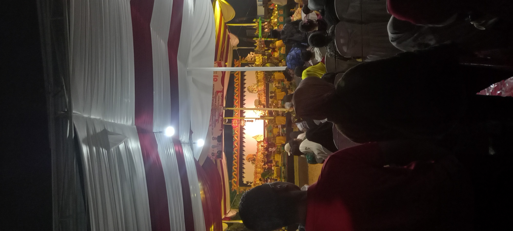 The image depicts an outdoor event, likely a cultural performance, taking place at night in Wanareja, Indonesia. The scene is illuminated by artificial lights, with a stage set up in the background featuring a screen on which shadow puppets are projected, characteristic of a Wayang Kulit performance. A banner above the stage reads "HUT KE-57 KEMERDEKAAN REPUBLIK INDONESIA TINGKAT KE CAMATAN WANGUREJA," indicating the event is celebrating the 57th Independence Day of the Republic of Indonesia at the sub-district level of Wanareja.
In the foreground, the backs of several attendees are visible, suggesting they are watching the performance. They are seated on chairs arranged in rows. The environment is covered by a large tent or canopy with red and white fabric draping, providing shelter and creating a festive atmosphere. The overall impression is of a community gathering for a traditional cultural event.