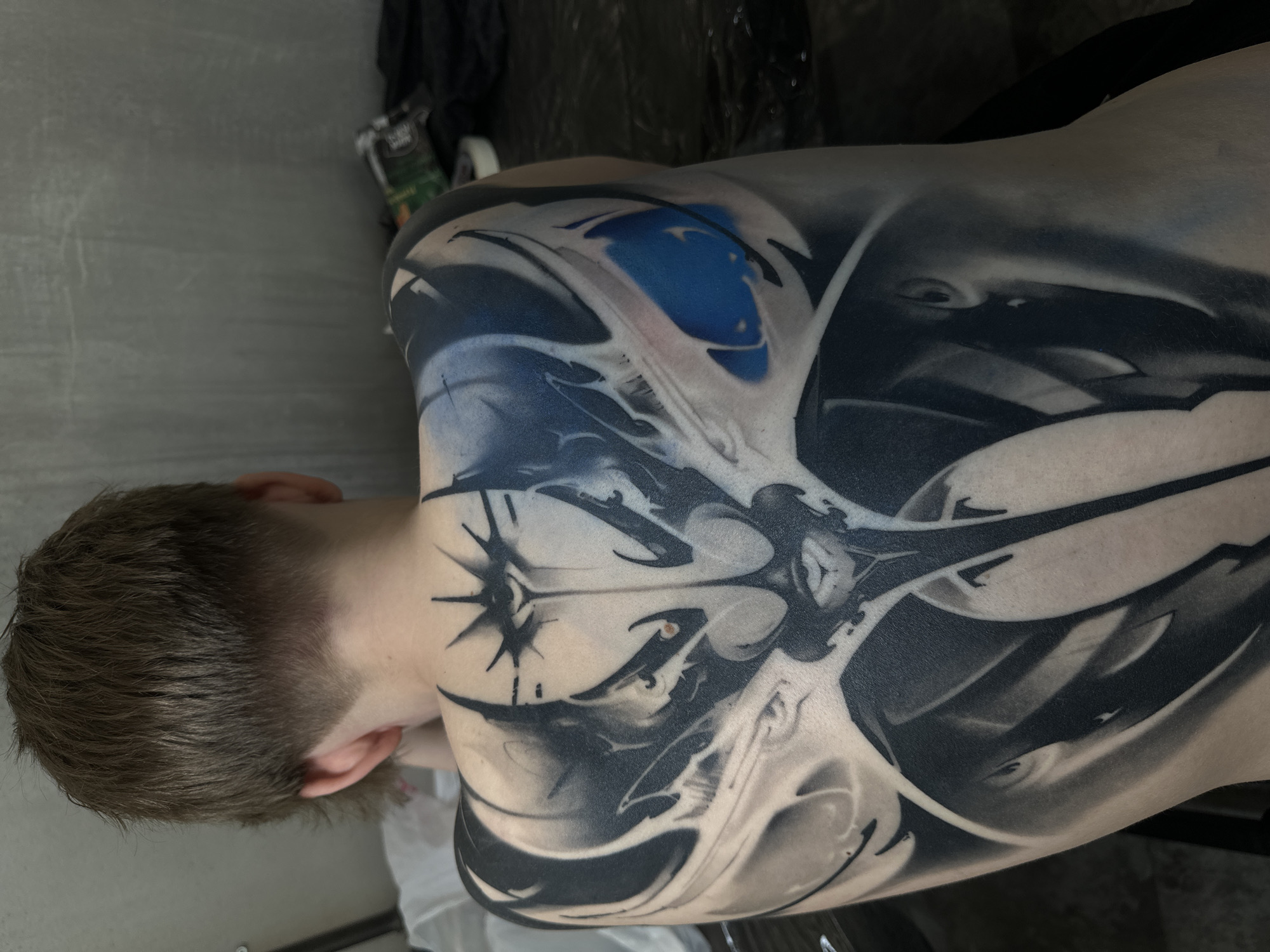 The image depicts a person's back with a large, intricate black and white tattoo that extends from the upper back and shoulder to the lower back area. The tattoo incorporates abstract shapes, lines, and some shaded areas, with a small section of blue coloring near the top. A stylized starburst is visible on the upper back near the neck. The person has short, light brown hair, and only the back of their head and part of their ear are visible. The background is out of focus, but it appears to be an indoor setting, possibly a tattoo studio or similar location, with some equipment and a dark-colored cloth or drape visible in the background. There is no location that is evident, and the city of Moscow cannot be verified.