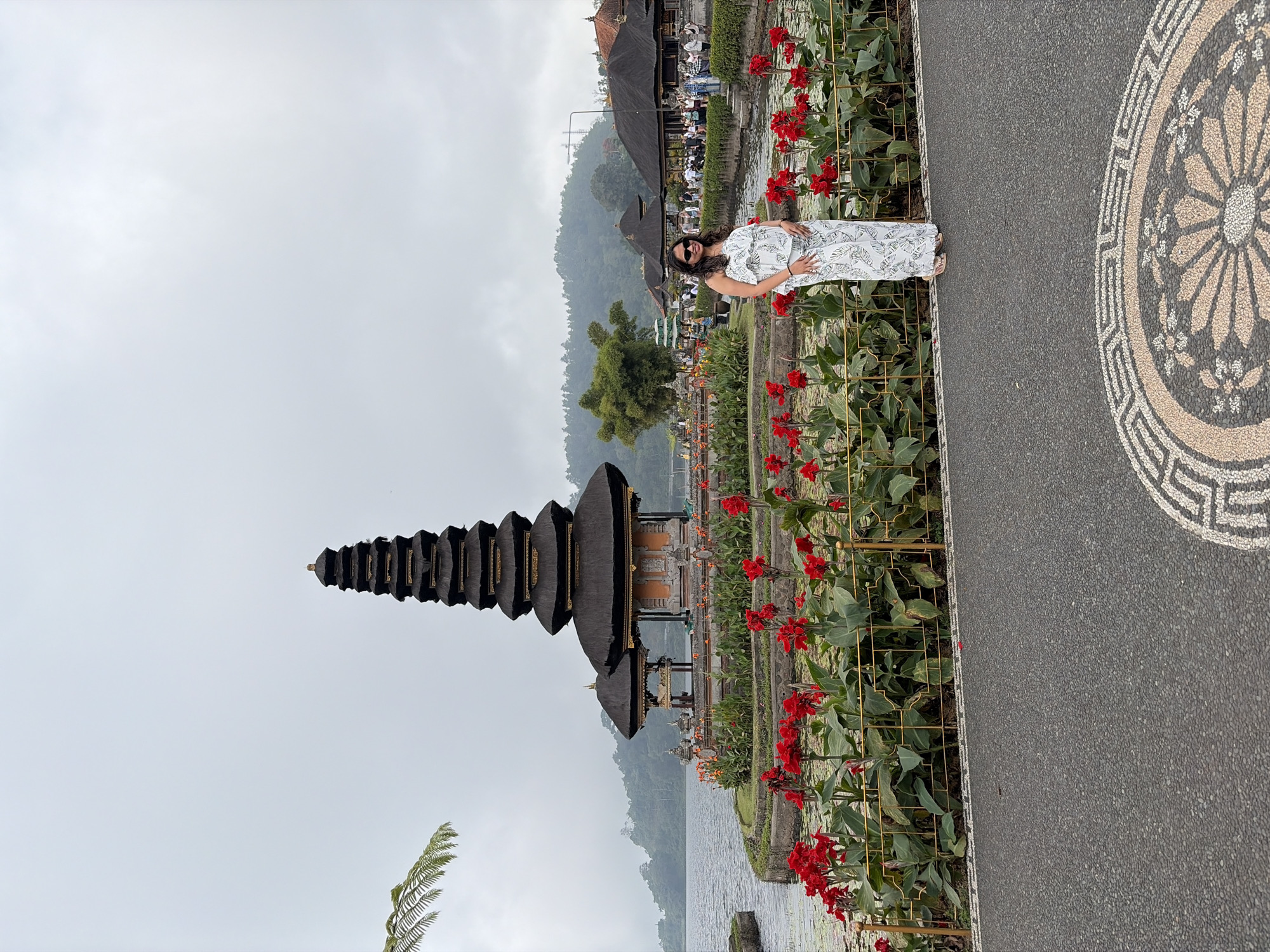 A woman stands on a paved pathway in front of a temple complex on a cloudy day. The woman is wearing a white, tropical-print jumpsuit and sandals. She is smiling and looking towards the viewer. Behind her, a multi-tiered temple with a dark, thatched roof rises from a body of water. The temple is surrounded by lush greenery and vibrant red flowers. In the background, there are other buildings with thatched roofs and a crowd of people. The location is Pura Ulun Danu Bratan, a Hindu temple in Baturiti, Indonesia, situated on the shores of Lake Bratan. The pathway in the foreground features a decorative mosaic pattern. The overall scene is serene and picturesque.