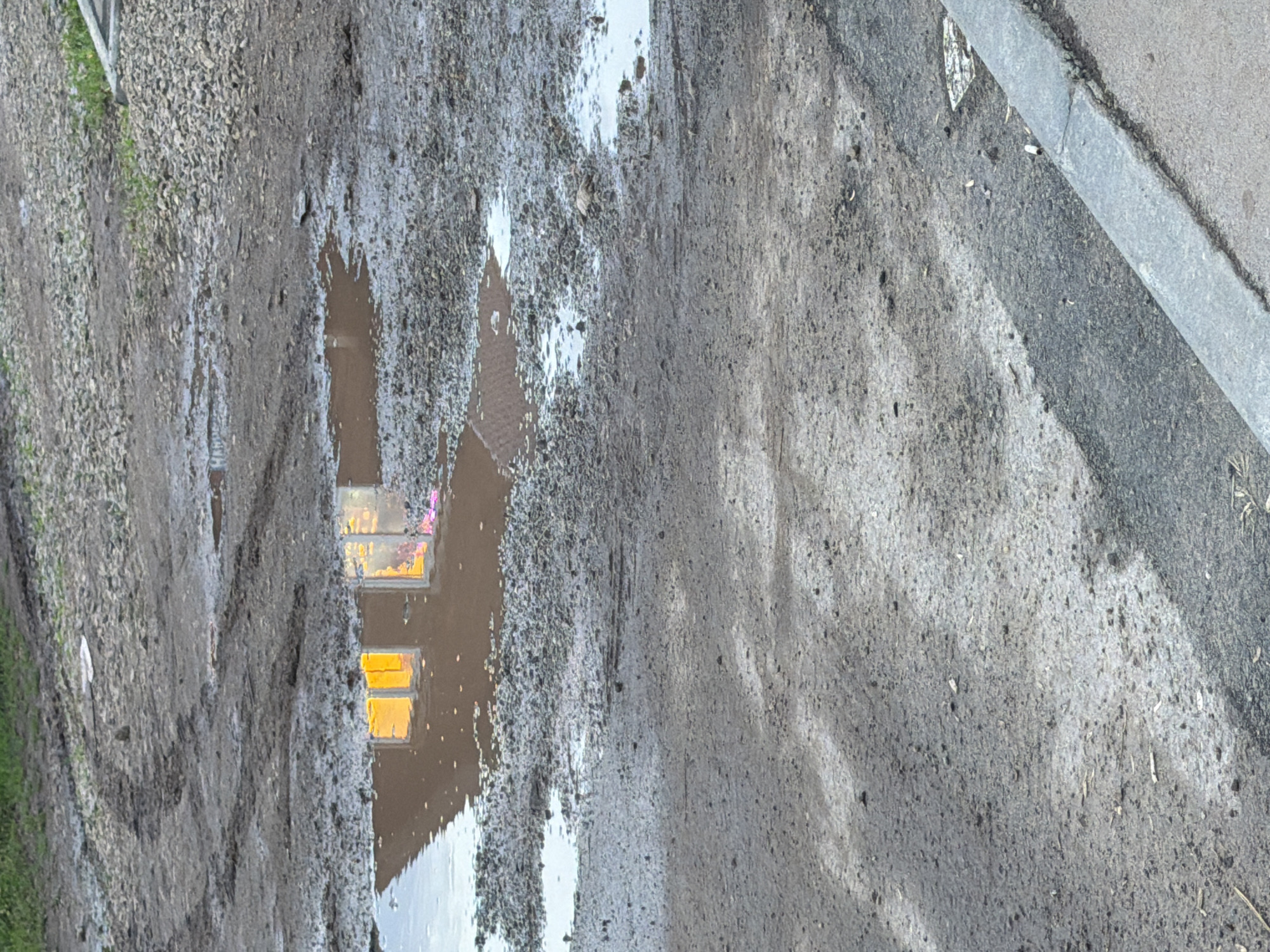 The image depicts a muddy, uneven dirt road with several puddles reflecting a building with illuminated windows. The puddles are located in the foreground and midground of the image, with tire tracks visible in the mud. In the background, there is gravel and some vegetation. The reflection shows two rectangular windows with bright yellow light emanating from them, suggesting interior lighting. The overall environment appears to be outdoors, likely in a rural or undeveloped area. The location is identified as the city of Froly, Russia.