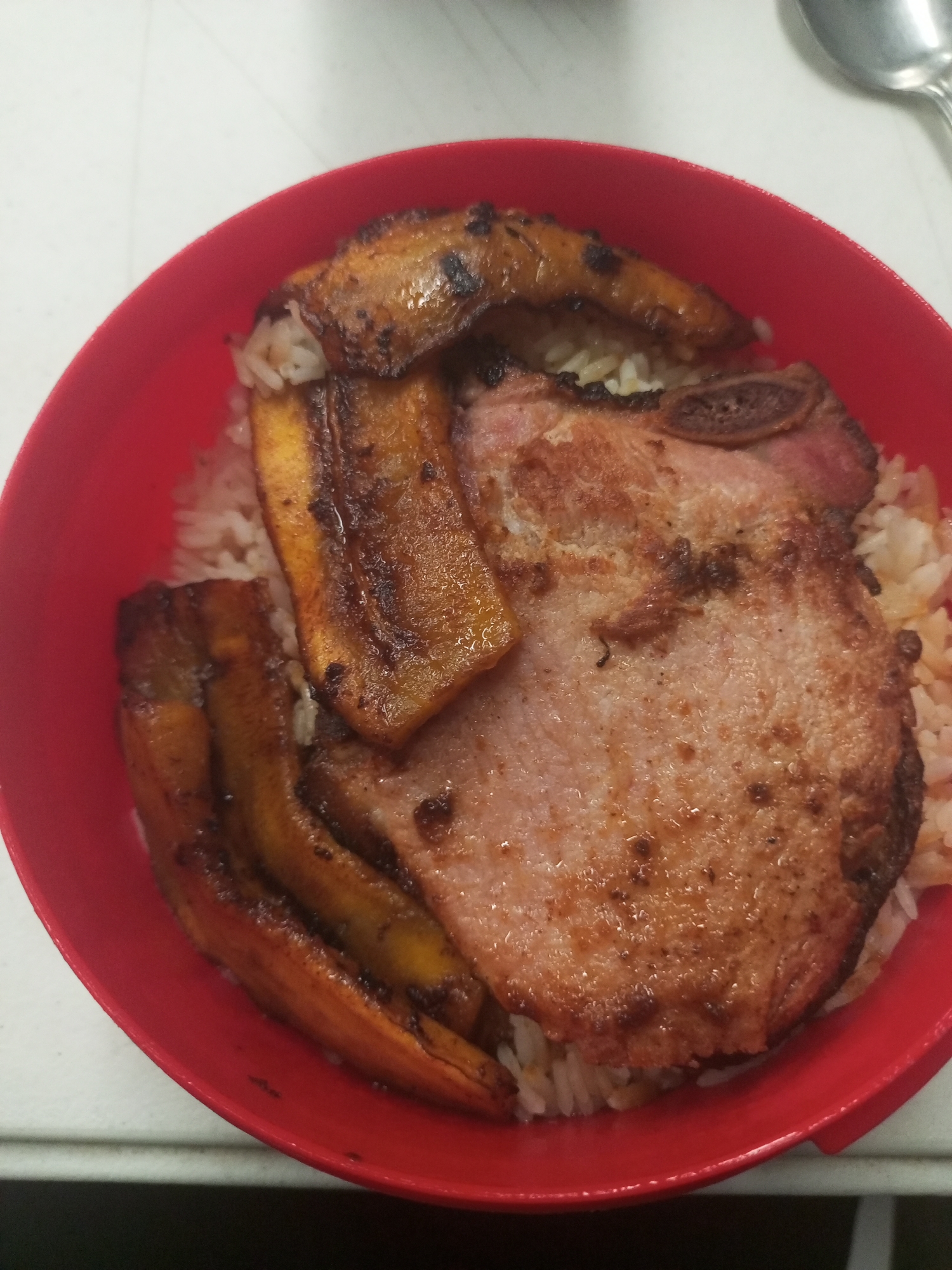 A red bowl contains a meal of white rice topped with a pork chop and fried plantains. A spoon is partially visible in the background.