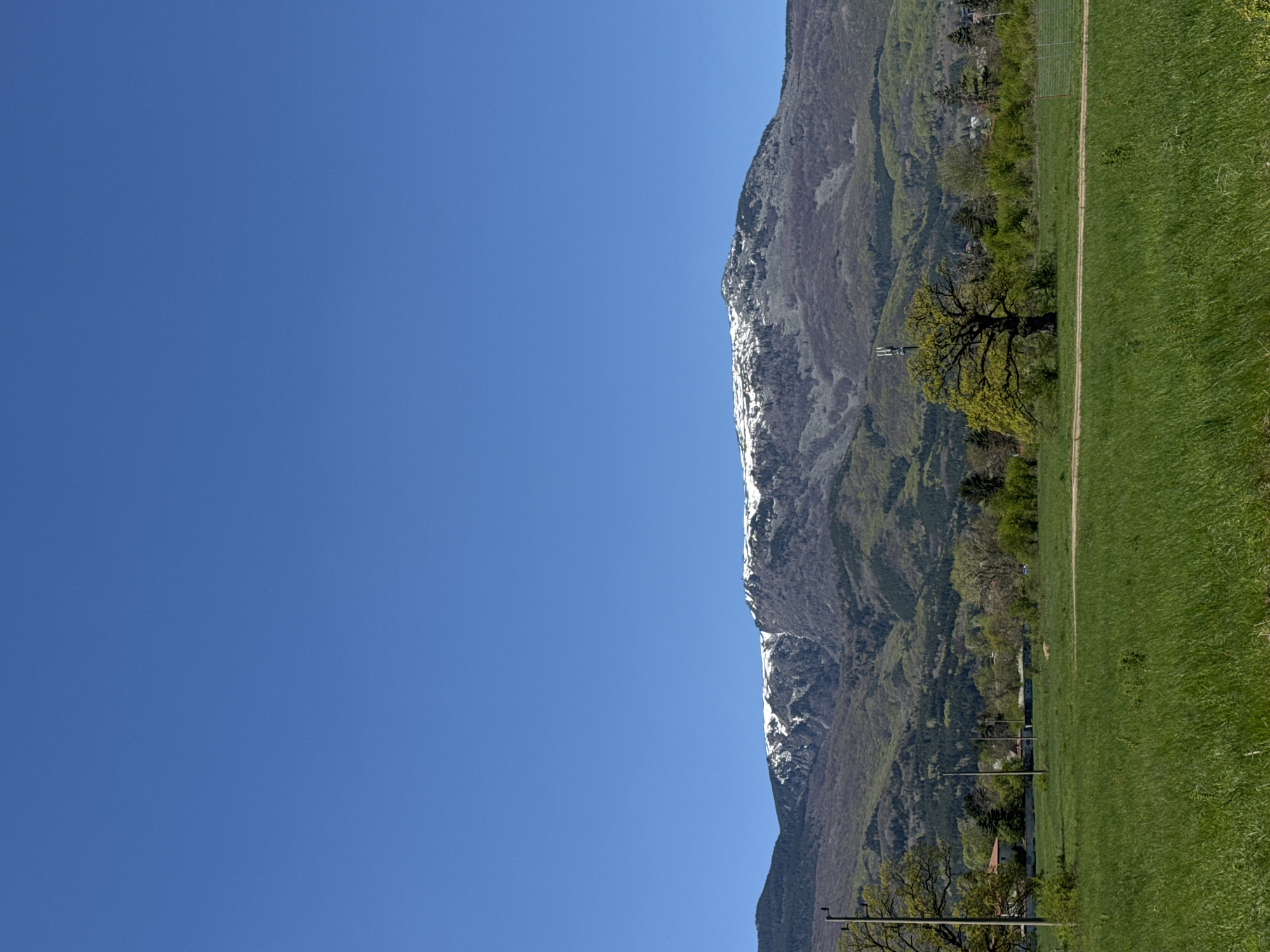 The image shows a mountain range partially covered in snow, bordering a green field with some trees. The sky is clear blue. The location appears to be a rural area, not within a city such as Sofia, Bulgaria. The absence of typically urban elements like buildings or dense infrastructure suggests a natural setting. There are no people visible. Power lines and possibly a few structures are at the base of the mountain.