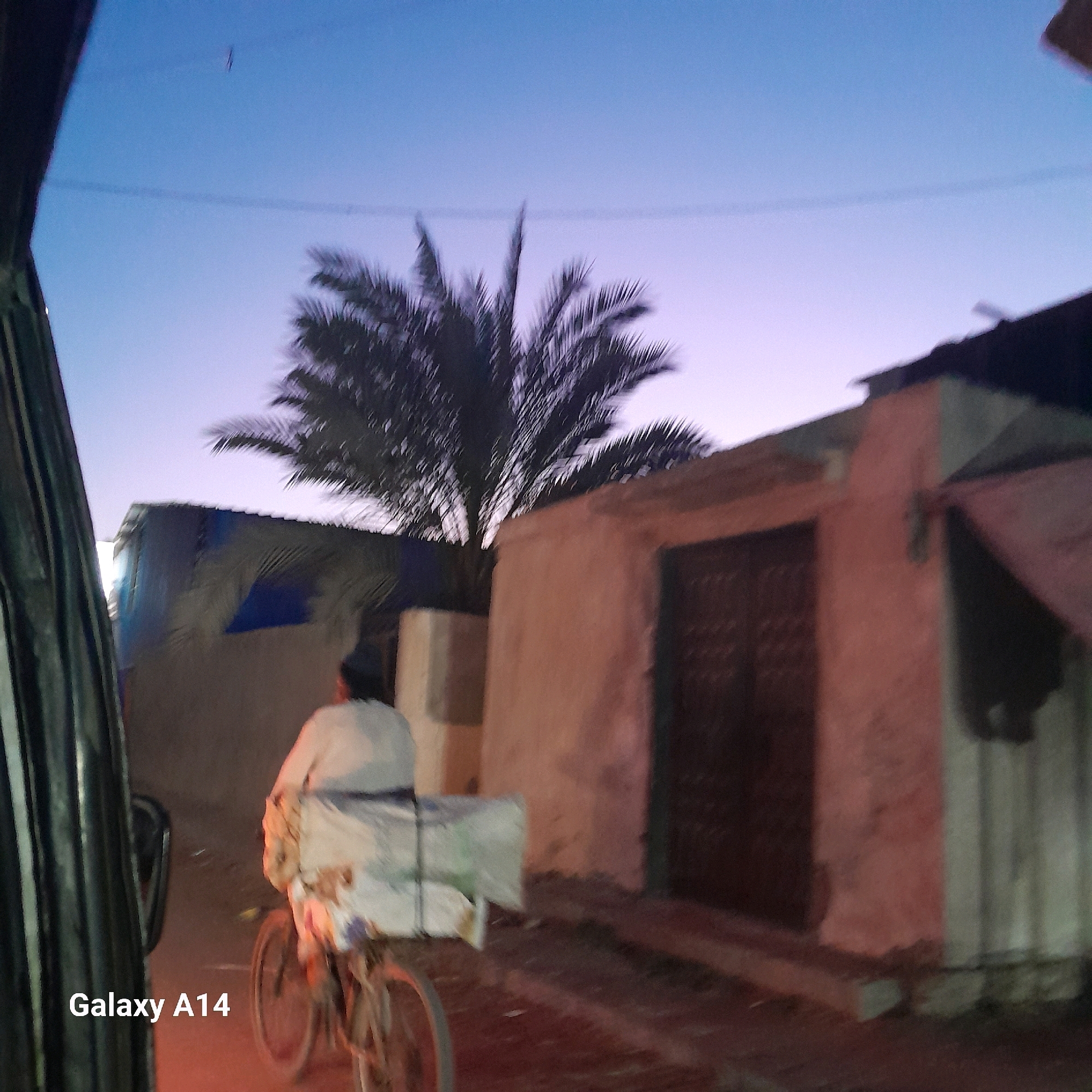 In Deir el-Balah, Palestinian Territories, the scene captures a dusky twilight sky. A person is riding a bicycle, carrying a large, rectangular package on the back, suggesting a delivery or transport of goods. The bicycle appears to be older, with visible spokes on the wheels. The rider is wearing a light-colored garment.
The environment includes a building with a peach-colored facade and a dark, possibly wooden, door. Next to it, another structure is visible, partially covered with what looks like a blue tarp or corrugated metal sheeting, with a darker roofline. A palm tree with sparse fronds is silhouetted against the sky. The foreground shows the left side of a vehicle, possibly a car or van, with a dark, textured interior panel. The ground is paved and shows some signs of wear and scattered debris. The "Galaxy A14" watermark is visible in the bottom left corner, indicating the device used to capture the image.