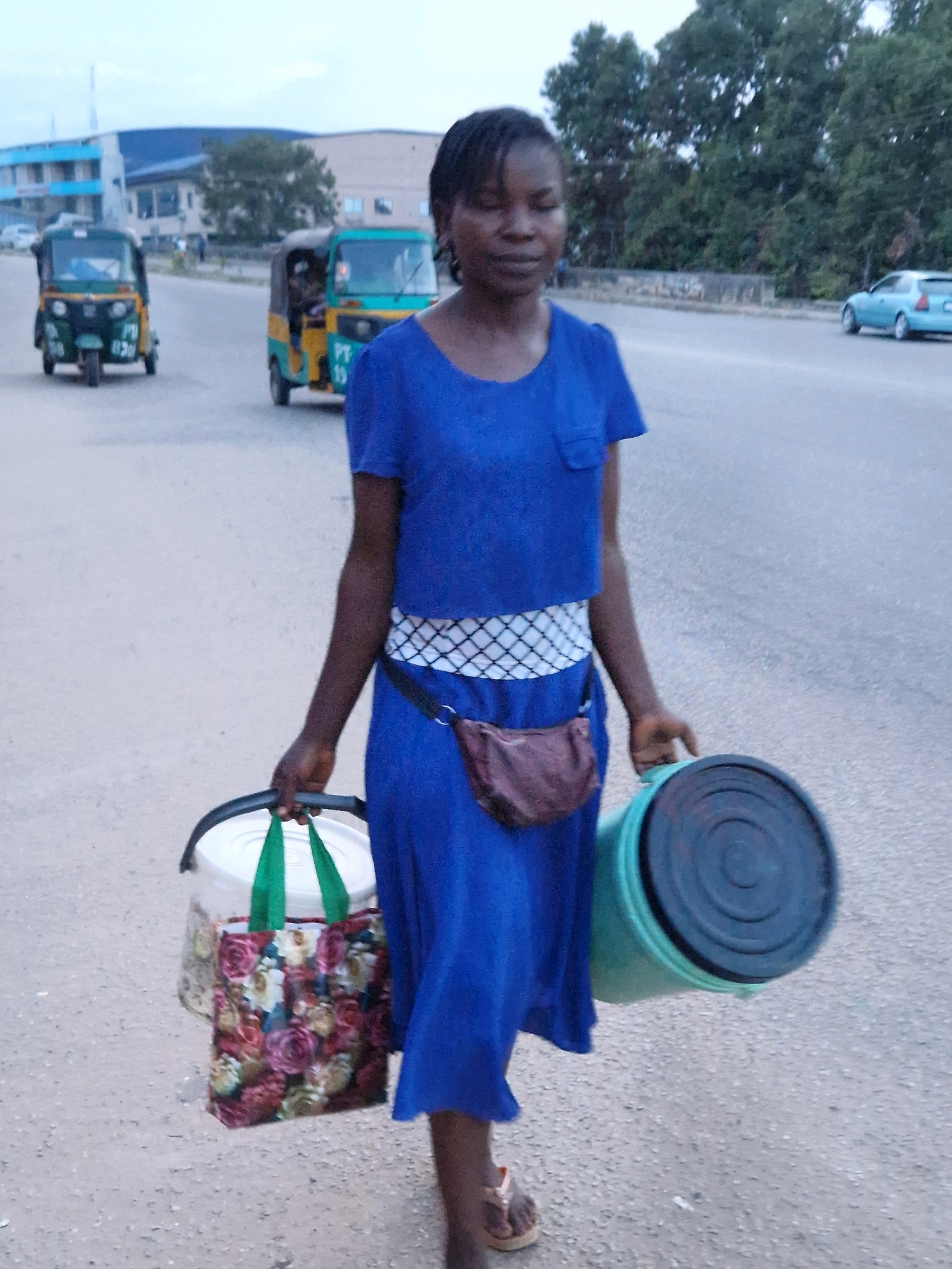 A young Black woman is walking on a paved road in what appears to be a city environment, possibly New Stadium, Nigeria. She is wearing a bright blue dress with a white and black patterned belt around her waist, and a brown fanny pack. She is carrying two items: a white bucket with a floral bag hanging from its handle on her left side, and a large, teal-colored plastic container with a black lid on her right side. The bucket on the left appears to contain some sort of items covered by the floral bag. She is wearing light brown sandals. In the background, several other vehicles are visible, including two green and yellow auto-rickshaws, and a light blue car. The road is asphalt and the surroundings include trees and buildings. The sky is overcast.