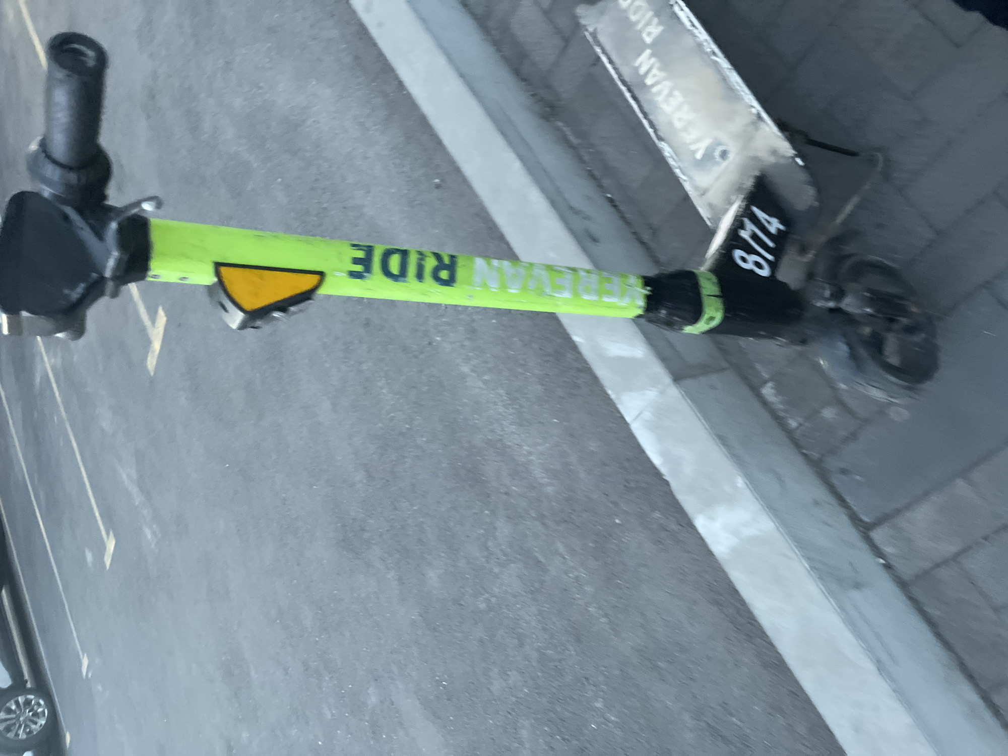 The image shows a Yerevan Ride electric scooter parked on a paved surface next to a concrete curb. The scooter is lime green with black components. The words "YEREVAN RIDE" are printed in large, capitalized letters on the scooter's stem. A registration number "8/14" is visible on the front of the scooter, near the front wheel. The background includes a gray asphalt surface with white painted lines, suggesting a parking area or street. The scooter is oriented at an angle in the frame, and the overall scene is brightly lit.