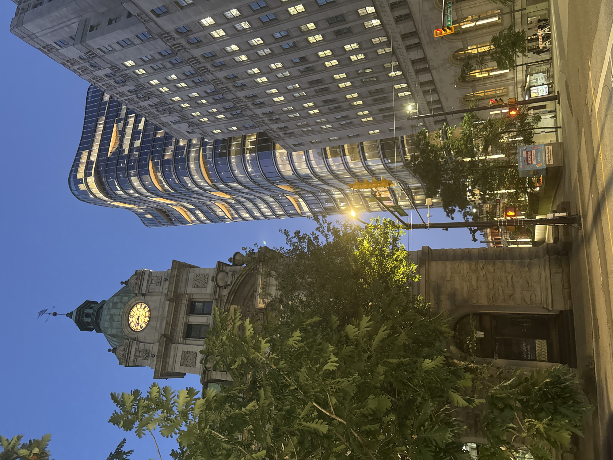 This is a vertical, full-shot photograph of a street scene in Vancouver, Canada, at dusk. The image is dominated by two prominent buildings: on the left, a historical stone building with a clock tower, and on the right, a modern, glass-facade skyscraper. The clock on the tower displays the time as approximately 6:15. Lush green trees partially obscure the lower portion of the stone building, their leaves illuminated by streetlights. The modern building features a curved design with many windows, some of which are lit, reflecting the twilight sky. The sky itself is a deep blue, indicating the transition from day to night. Traffic lights are visible, one red and one yellow, along with street signs for W Hastings St. Several saplings and larger trees are planted along the sidewalk. In the foreground, a street is visible with white lane markings. The scene appears to be a busy intersection in a downtown urban environment.
