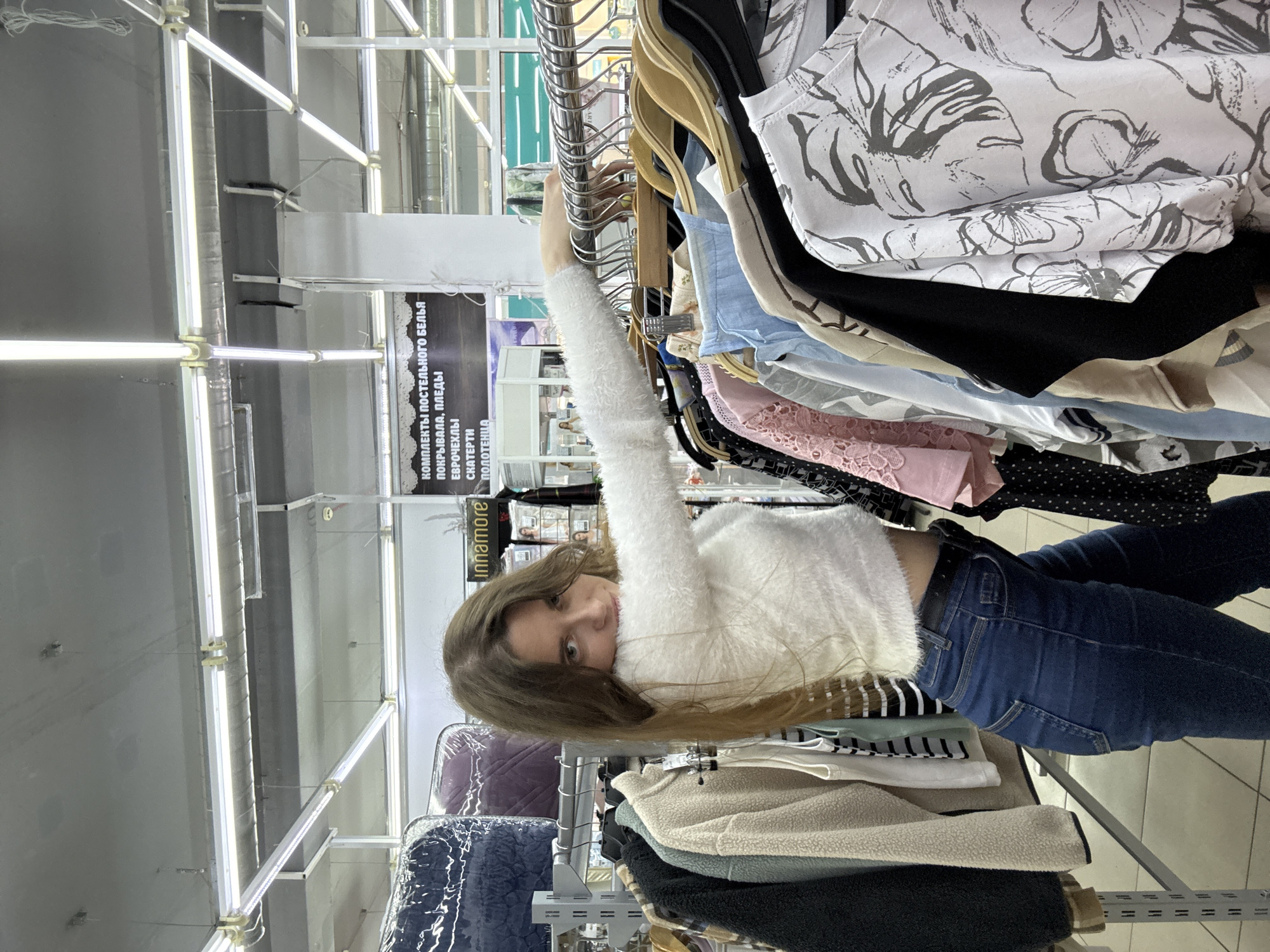 A woman is browsing clothes on a rack in a store in Chaikovsky, Russia. She is wearing a white fuzzy sweater and blue jeans. The clothes on the rack include a variety of colors and patterns. The store has fluorescent lighting. A sign in the background includes Russian text, advertising bedding and towels.