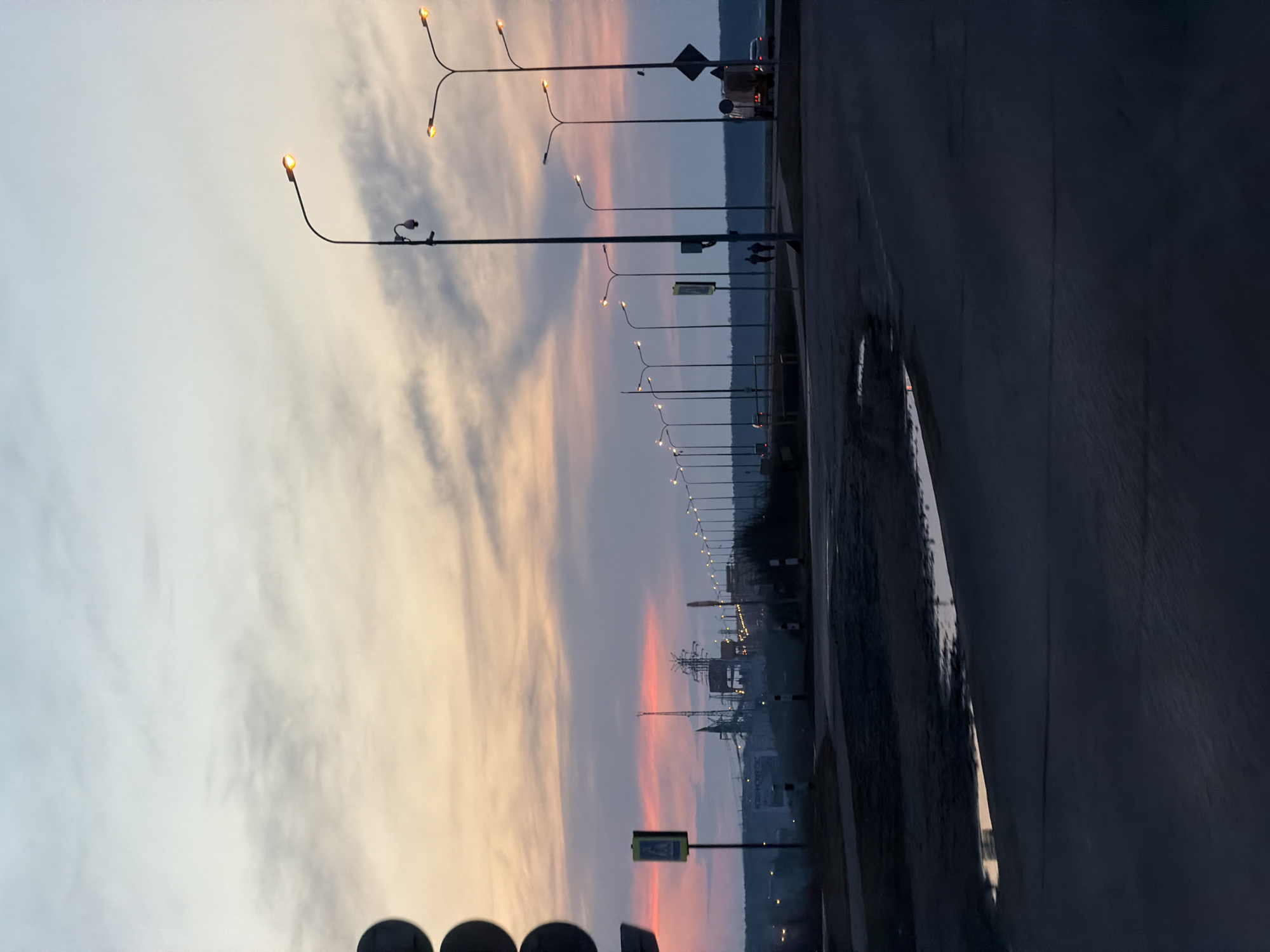 The image depicts a street scene in Chaikovsky, Russia, at dusk. A line of streetlights illuminates the road, their warm glow contrasting with the cool tones of the sky and the darkening landscape. The sky displays a gradient of colors, ranging from light blue to orange and pink. An industrial complex with towers and structures is visible in the distance, silhouetted against the vibrant sky. The road surface is wet, reflecting the surrounding light. A pedestrian crossing sign is visible along with blurred shapes in the foreground. Two people are walking on the side of the road further into the image. The angle of the picture suggests a perspective from either within a car, or with a tilted camera.