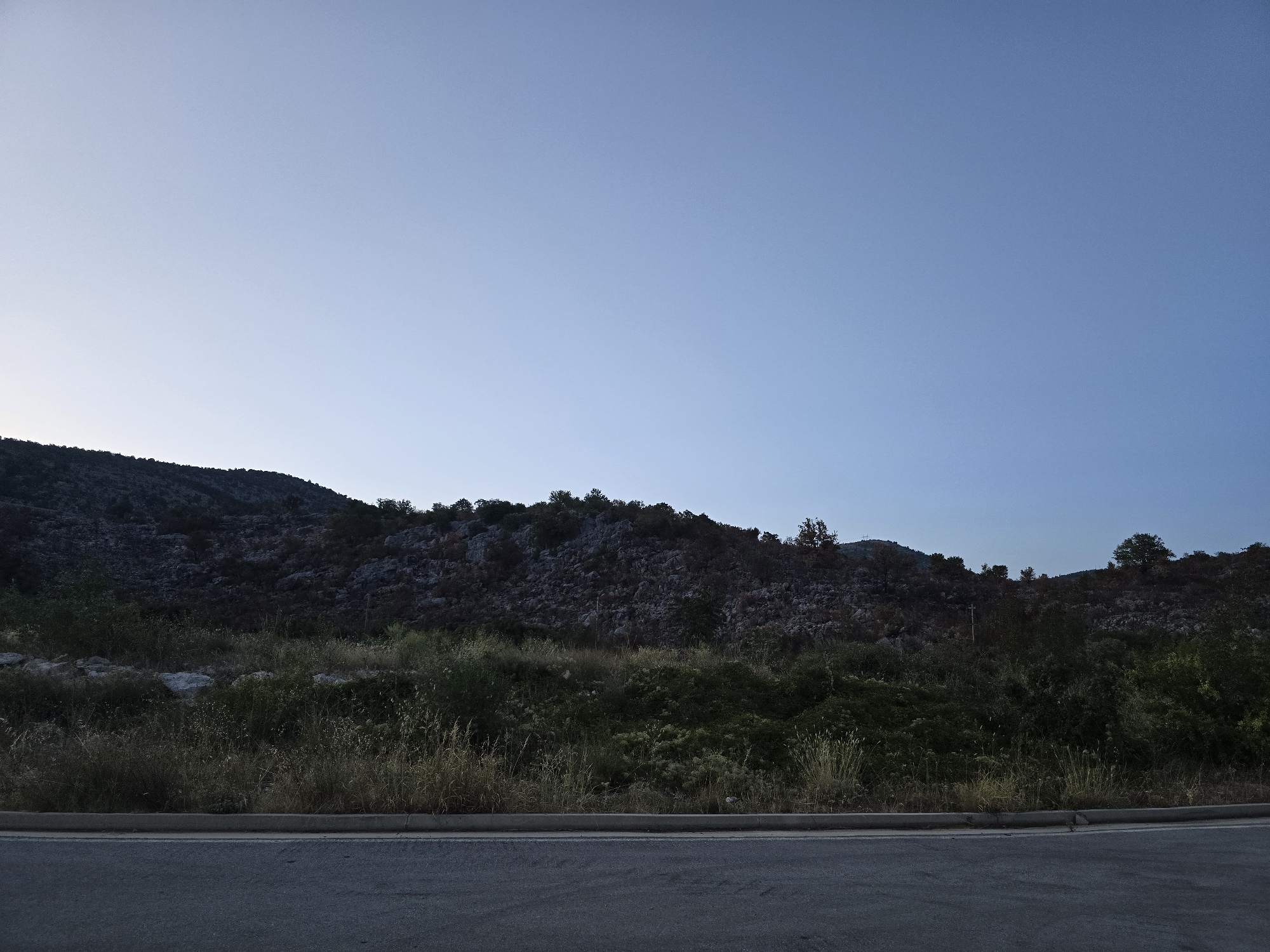The image depicts a roadside scene at the base of a rocky hillside, likely in Klopot, Montenegro, under a clear, pale blue sky. In the foreground, a portion of an asphalt road is visible, with a concrete curb separating it from a band of dry, tall grass and various green vegetation. The hillside behind the vegetation is covered in dark, rough-textured rocks and sparse trees, with a few larger trees dotting the top edge of the hill. The lighting suggests it is either early morning or late evening, with a soft, diffused light casting subtle shadows. There are no people or animals visible in the frame.