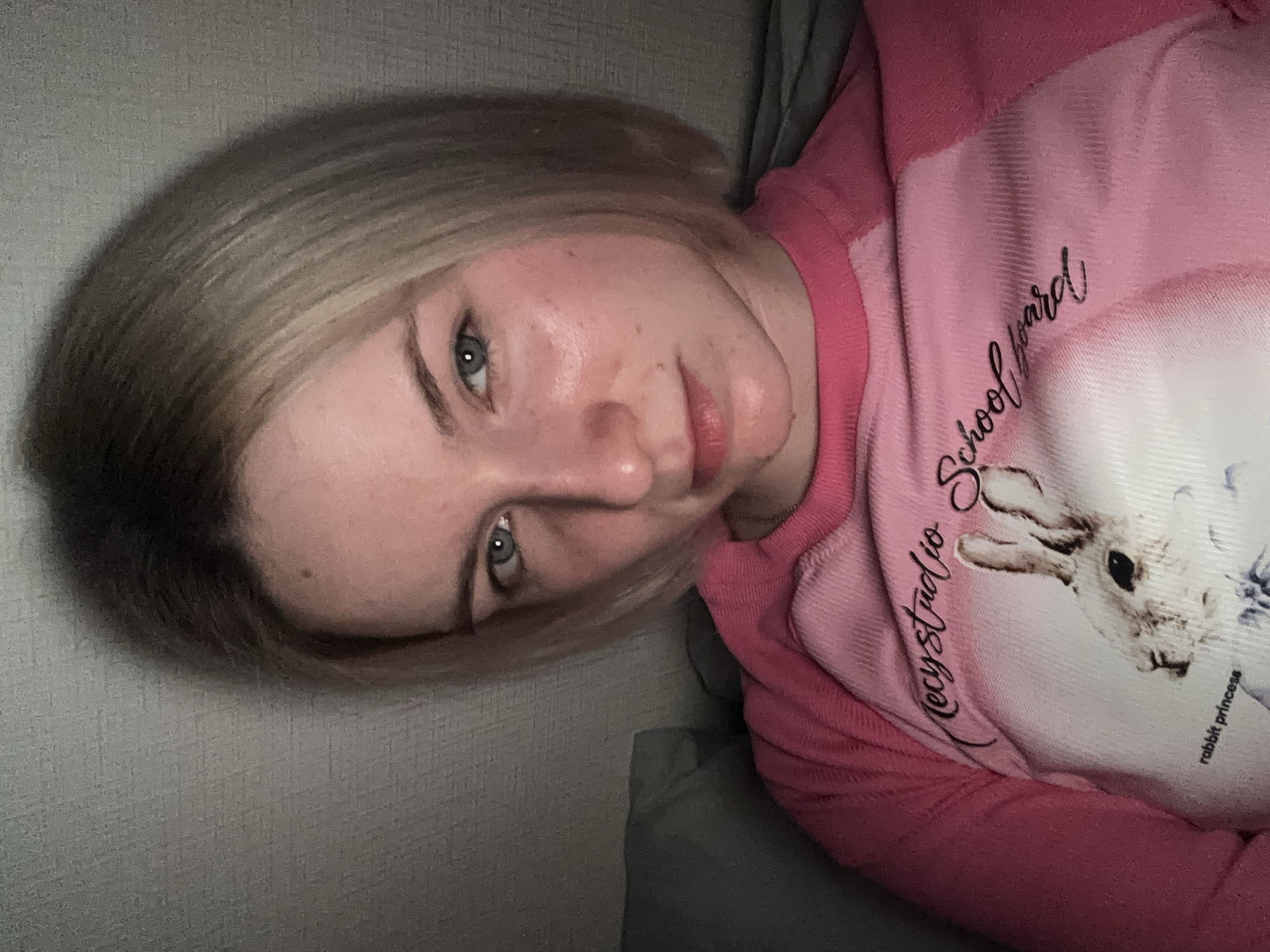 A medium-shot portrait depicts a young individual, likely female, lying down indoors. The person has short, light blonde hair with visible darker roots and light blue-green eyes, looking directly at the camera with a neutral to slightly contemplative expression. They are wearing a two-tone long-sleeved top, primarily pink, with a lighter pink or white central panel.
The top features a graphic of a white rabbit. Several lines of text are visible on the garment: "Mecy studio School yard" is written in a cursive, dark script above and to the left of the rabbit graphic, and "rabbit princess" is printed in smaller, dark text below the rabbit.
The background consists of a textured grey wall and what appears to be grey bedding or upholstery, suggesting a casual indoor environment such as a bedroom or living room. The lighting is dim and low-key, indicating an evening or night time setting. The overall mood of the image is relaxed and informal. The scene is located in Ligolambi, Russia.