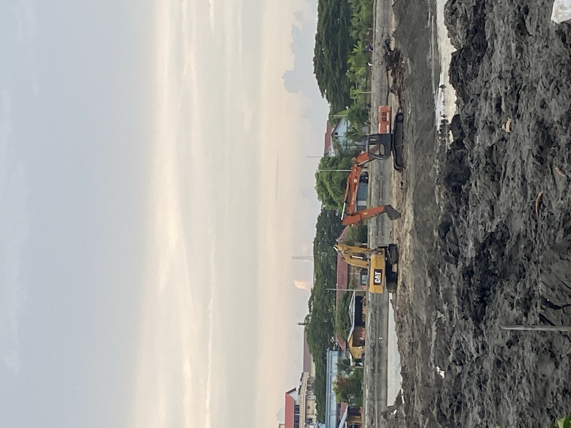 Two excavators, one yellow CAT and one orange Hitachi, are engaged in land reclamation work at a construction site in Lhokseumawe, Indonesia. The CAT excavator is positioned to the left of the frame, while the Hitachi is to the right. The ground is characterized by mud and debris, indicating ongoing excavation and earthmoving. In the background, buildings and trees suggest an urban or semi-urban environment. The sky is overcast, with soft clouds dominating the upper portion of the image. Several individuals are visible near the water's edge, possibly workers or bystanders. The overall scene depicts an active construction project.