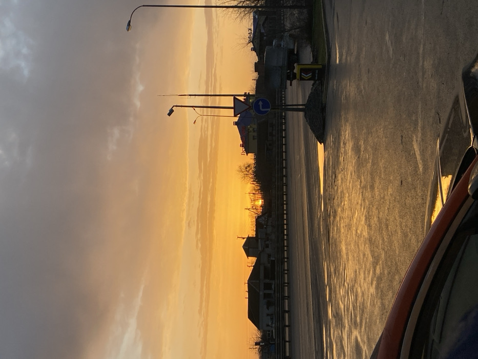 A sunset casts an orange glow across a street and sky. The scene includes streetlights, road signs, and buildings silhouetted against the vibrant sky. The road is wet, reflecting the warm light. A portion of a car is visible in the foreground, adding to the perspective of the street. The sky blends from gray clouds to bright orange near the horizon.