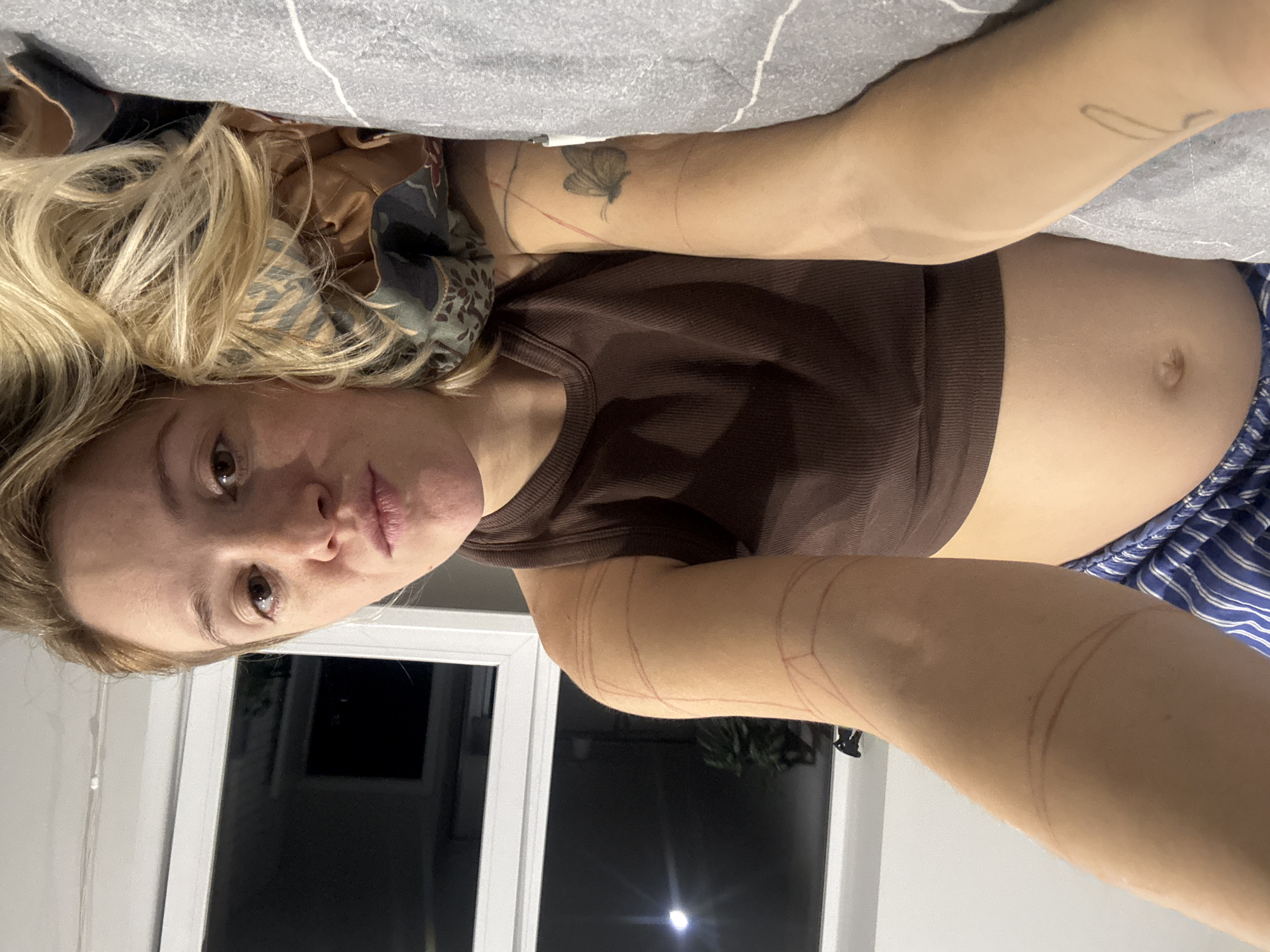 The media file contains a close-up, horizontal portrait of a woman lying on a bed. She has blonde hair and is looking towards the camera with a neutral expression. She is wearing a brown tank top and blue and white striped pajama bottoms. The room appears to be a bedroom, with a grey blanket visible in the background. A window with white blinds is also visible, showing a dark, nighttime scene outside. There are no visible text elements in the image. The overall mood is casual and intimate.