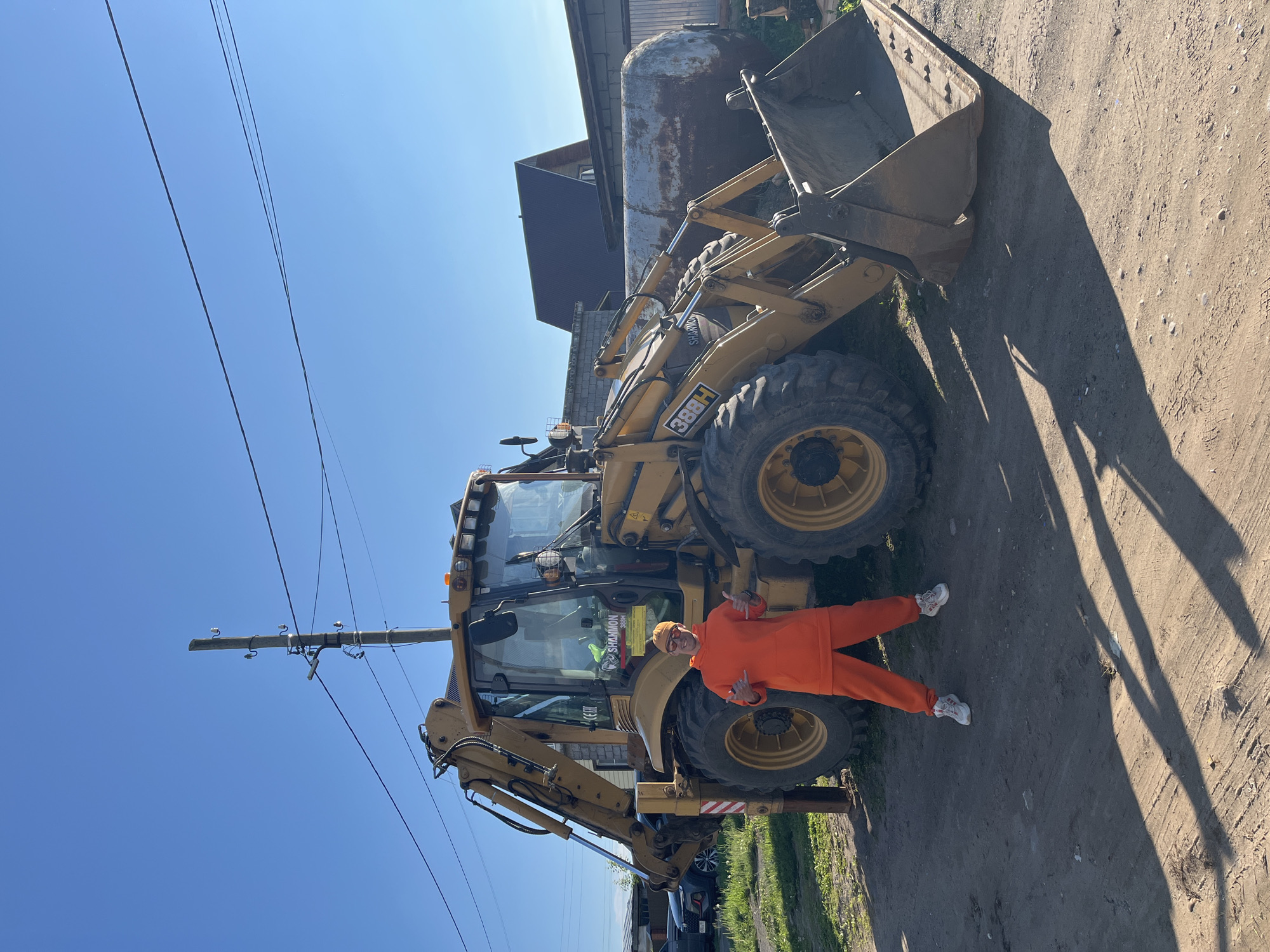 The image displays a full-length shot of a person standing beside a large yellow backhoe loader on a bright, sunny day in Chaikovsky, Russia. The person, appearing to be male, is dressed in a matching orange tracksuit, an orange beanie, and white sneakers. He is positioned to the right of the backhoe's front wheel, facing the camera with a cheerful expression and making "shaka" (hang loose) hand gestures with both hands.
The heavy equipment is a yellow backhoe loader, identified by "CAT 388H" and a sticker reading "SHAMMON 388H" on its side. It features large, dark-colored tires and a front bucket. In the background, under a clear blue sky, there's a large, rusty metal storage tank and several buildings, suggesting an industrial or rural setting. A utility pole with power lines runs vertically on the left side of the frame. The ground is unpaved dirt and gravel, showing tire tracks and sparse grass. Strong shadows cast by the machinery and the person indicate a sunny, likely mid-day to early afternoon light. The scene captures a casual moment with the heavy machinery.