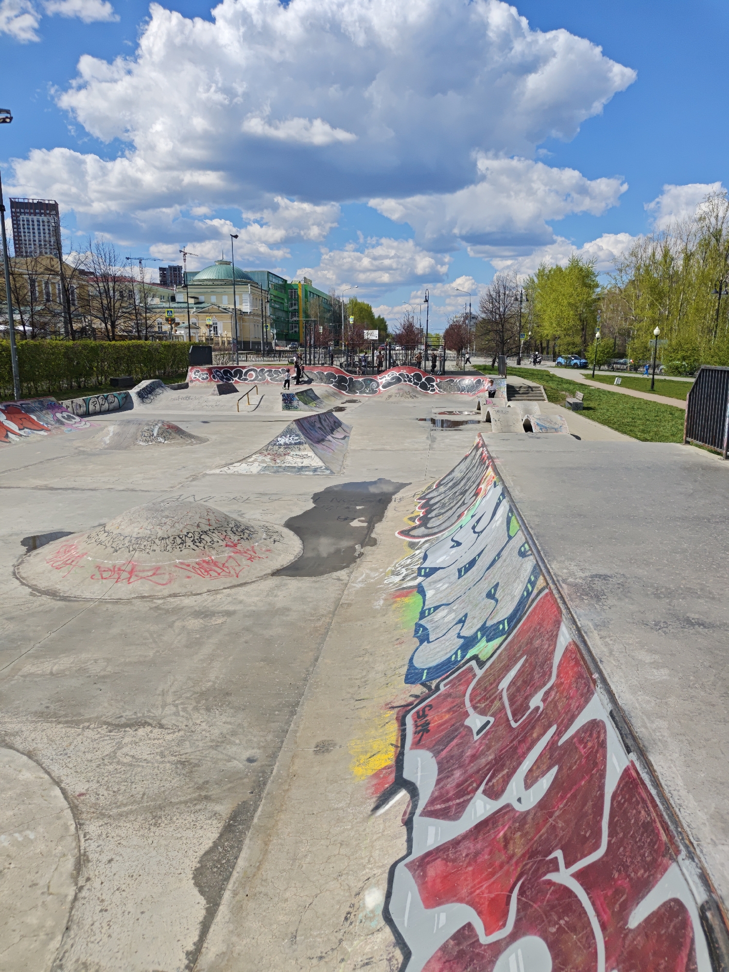 The image shows a skate park in Yekaterinburg, Russia. The park is made of concrete and features various ramps, bowls, and obstacles. Graffiti covers many of the surfaces. In the background, there are buildings and trees, and people walking along a sidewalk. The sky is blue with fluffy white clouds.