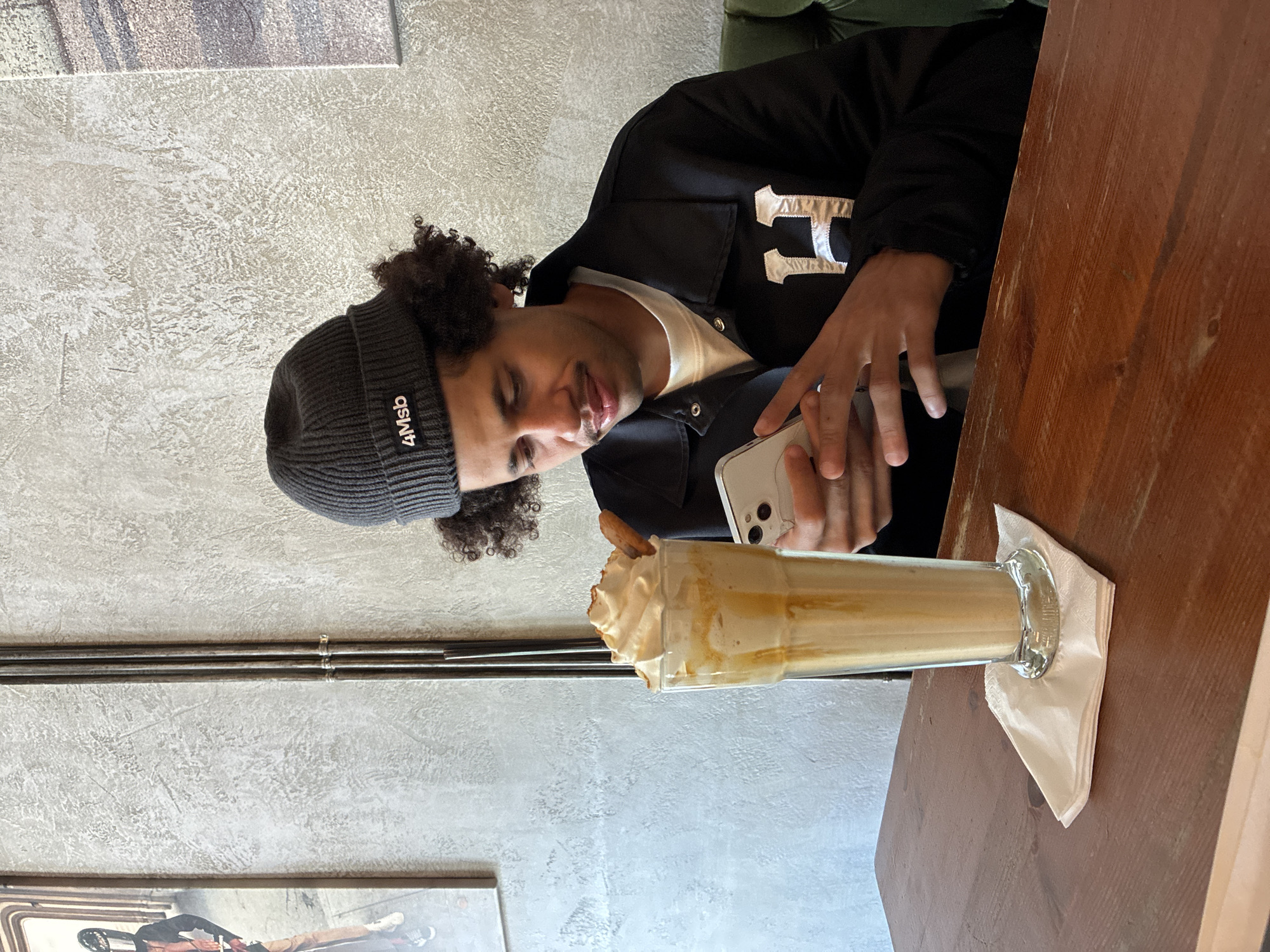 A young man with curly hair and a mustache is seated at a wooden table, looking down at his phone. He is wearing a dark gray beanie with "4Msb" written on it and a black jacket with a large white "H" on the chest. In front of him is a tall glass of a light brown, creamy beverage topped with whipped cream and a cookie. The scene appears to be indoors, possibly a cafe, with a textured gray wall and some framed artwork in the background. The lighting is natural and soft, casting a gentle glow on the man and his drink.