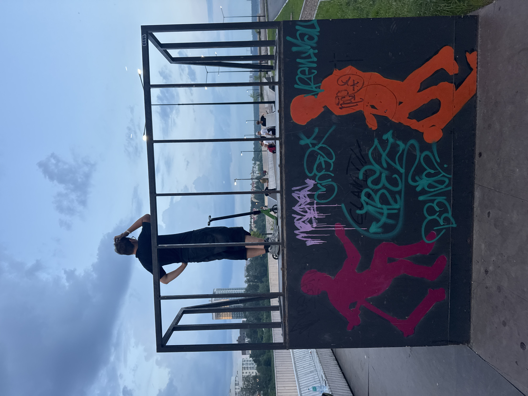 An overcast sky looms above a youth skatepark in Yekaterinburg, Russia. A young man, viewed from behind, stands at the edge of a raised platform, his hand on his head as if contemplating. He wears a black t-shirt and dark shorts. To his right, a green electric scooter rests against a black metal railing. The graffiti-covered wall of the skatepark features bold pink and orange silhouettes of figures, one appearing to ride a scooter, accompanied by spray-painted tags in purple and green. In the background, streetlights illuminate a pathway and distant buildings, suggesting an urban setting. A body of water, likely a river, is visible in the distance.